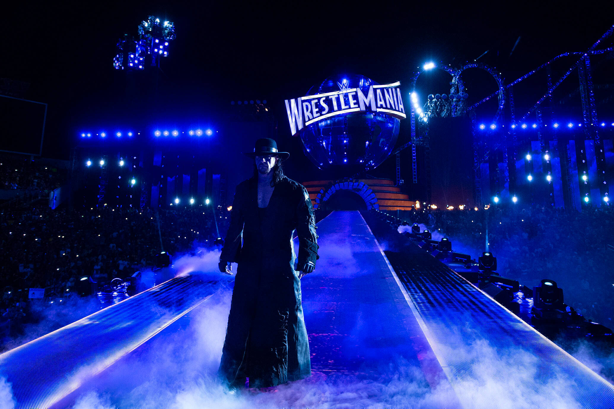 送葬者於摔角狂熱賽 壁紙 — The Undertaker 沿著藍色燈光的 WrestleMania 斜坡走下來，周圍環繞著霧氣和體育場燈光。