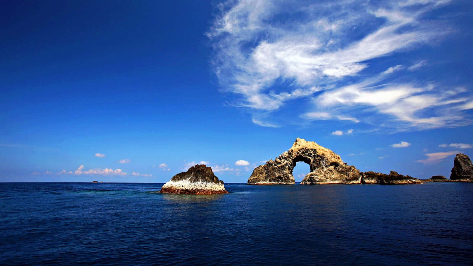 「逃離現實，享受高解析度海灘美景」 壁紙 — 一座雄偉的自然岩石拱門矗立在深藍色的海洋水域中，上方是充滿活力的藍天和縷縷白雲。