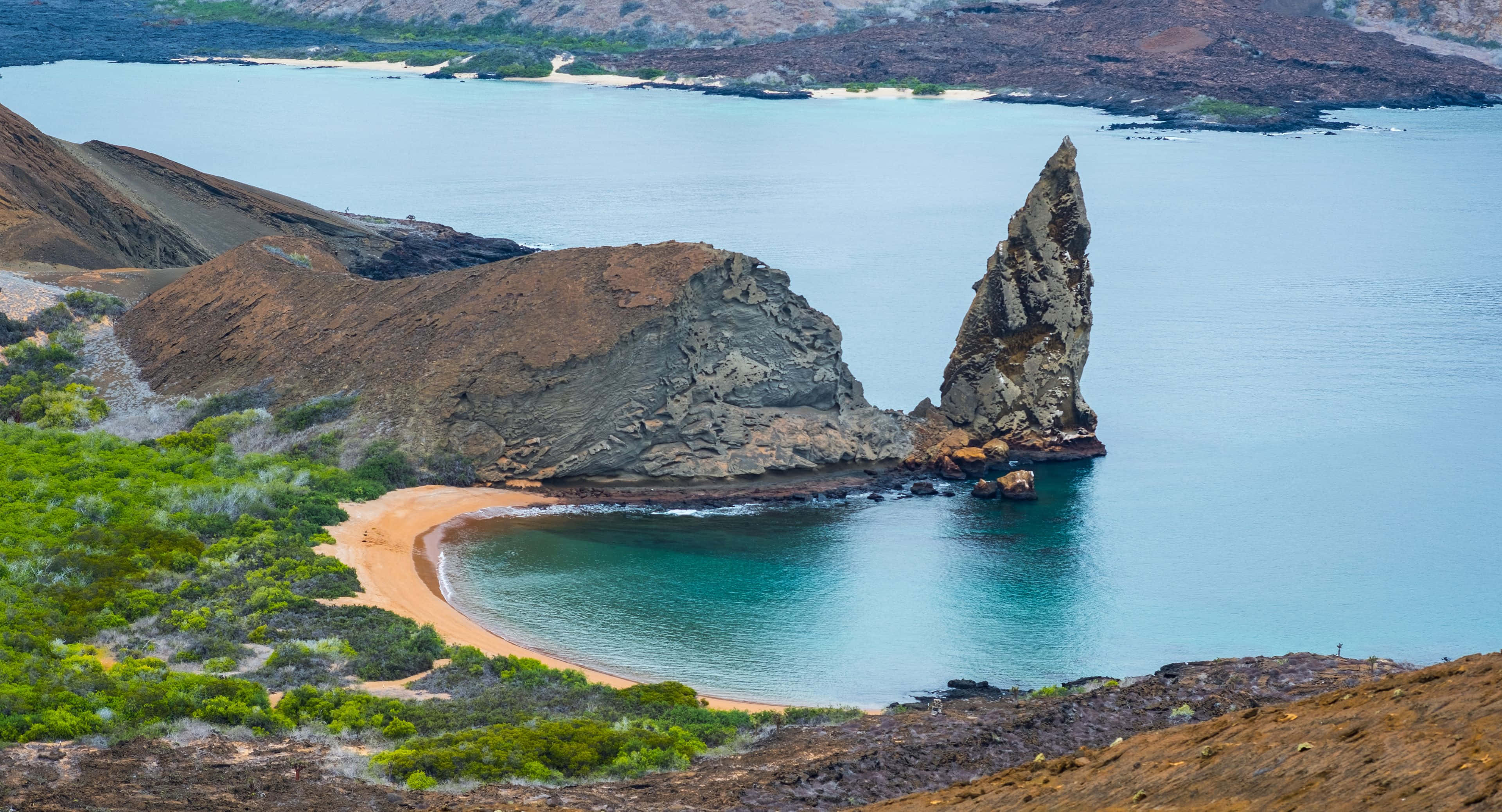 逆光沙漠風景 壁紙 — 一座孤獨高聳的岩石尖塔從碧綠色的海水中升起，旁邊是新月形沙灘，周圍環繞著崎嶇的棕色懸崖和綠色灌木。