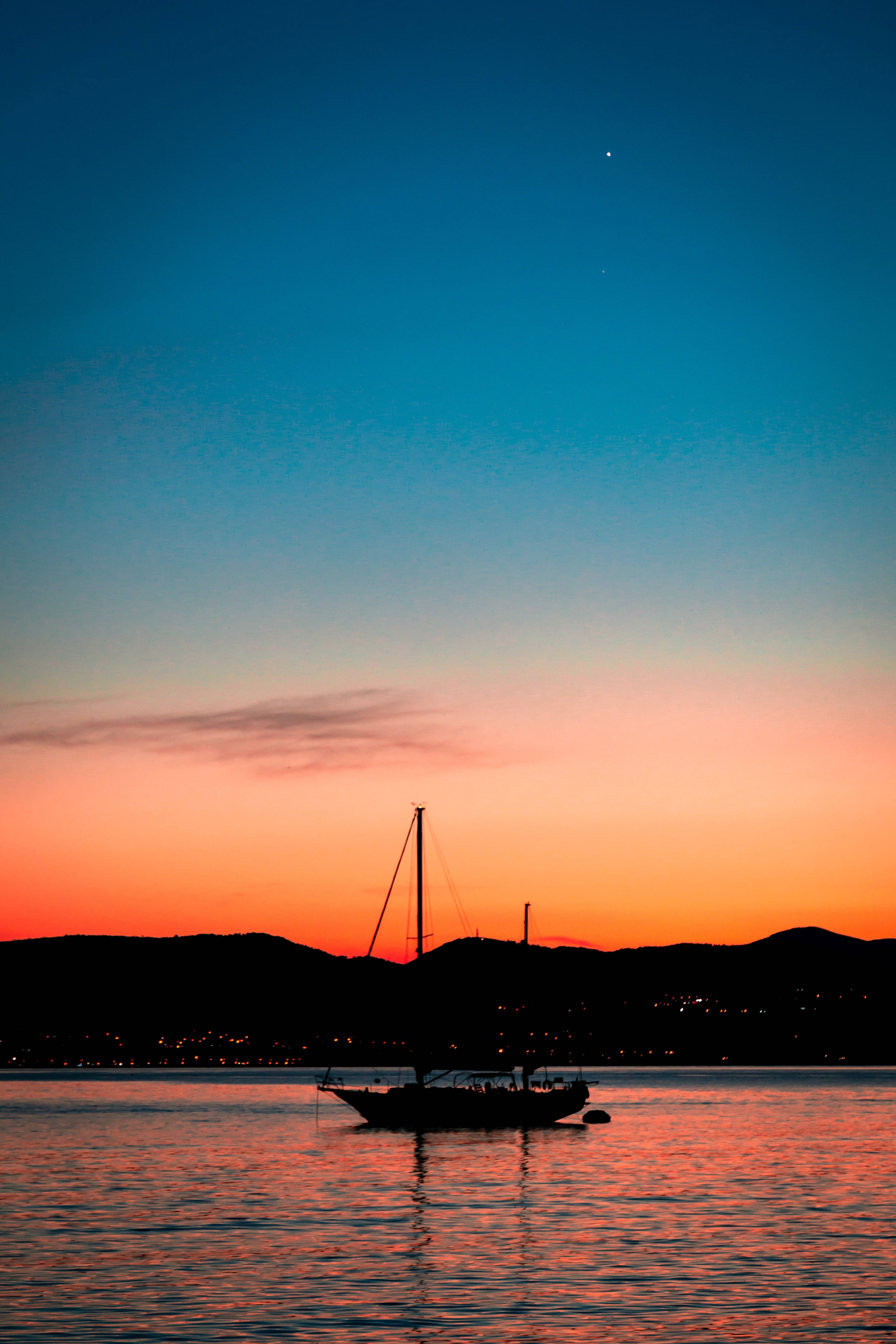 逆著絢麗暮色天空航行 壁紙 — 帆船剪影在平靜的水面上滑行，夕陽西下，遠山之上是充滿活力的橙色和藍色天空。