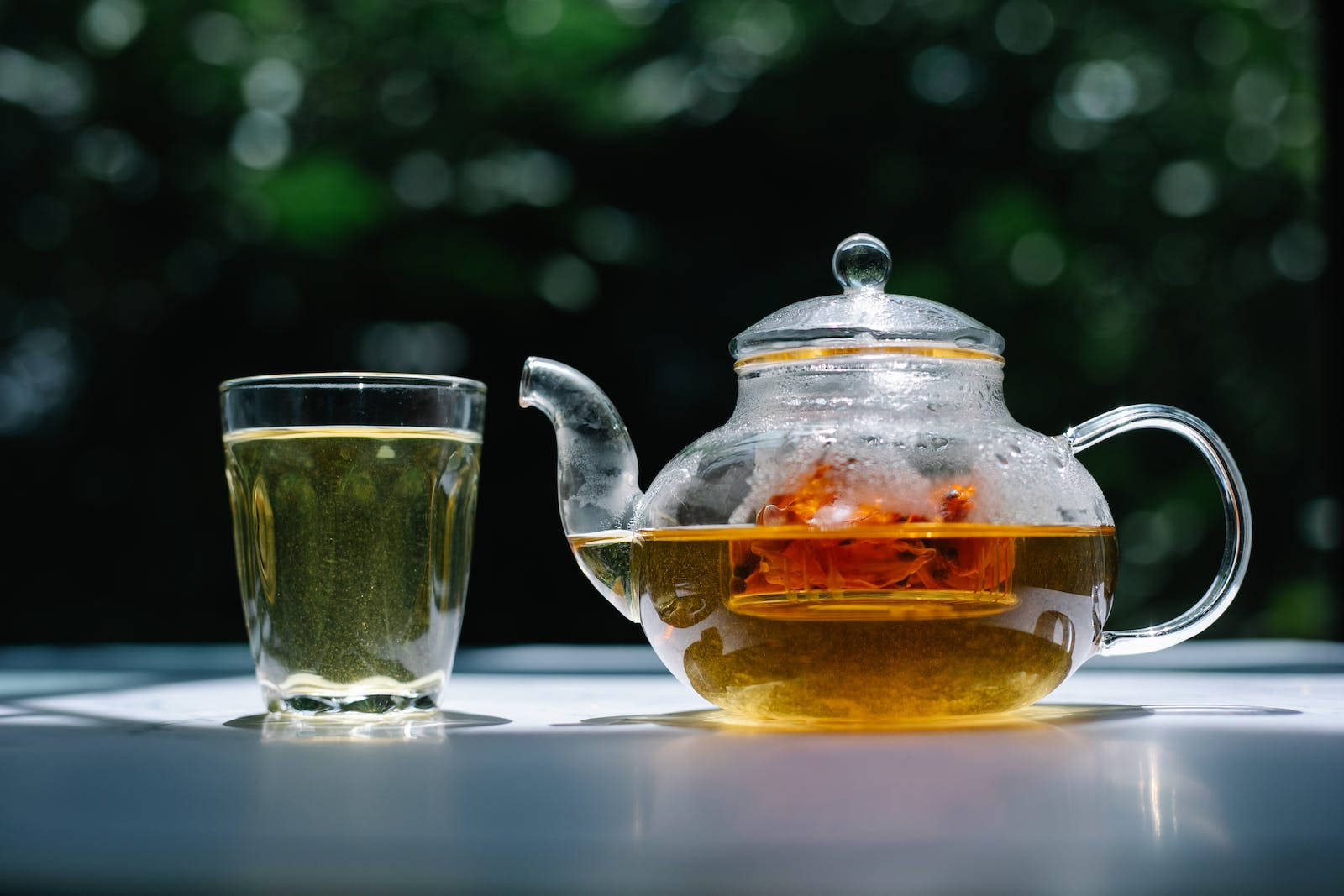 白色桌子上的玻璃茶壺和茶杯，背景是綠色樹葉。