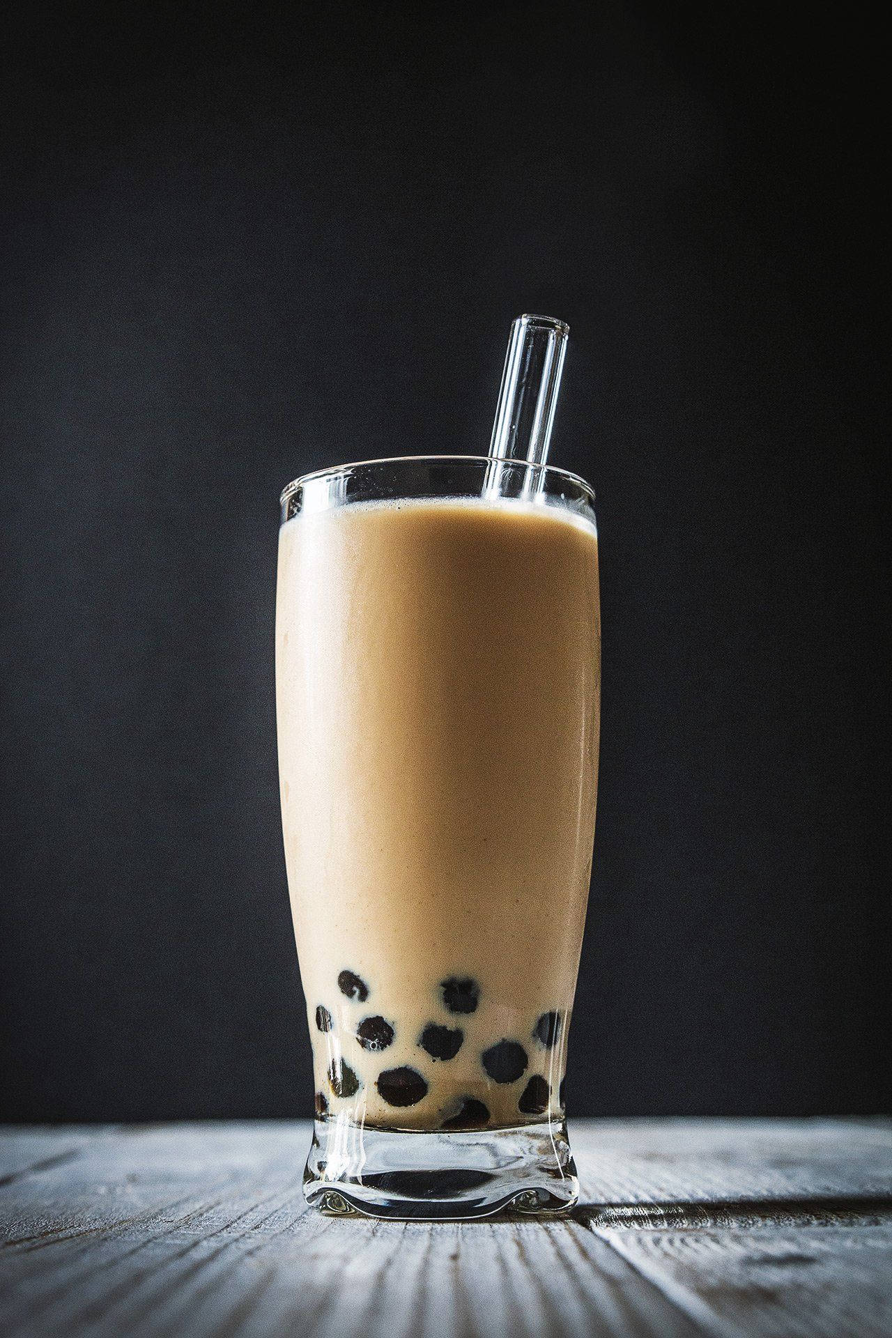 透明玻璃杯珍珠奶茶 壁紙 — 一杯高身嘅忌廉泡泡茶配黑色木薯珍珠，放喺深色背景嘅鄉村風格木枱上面。