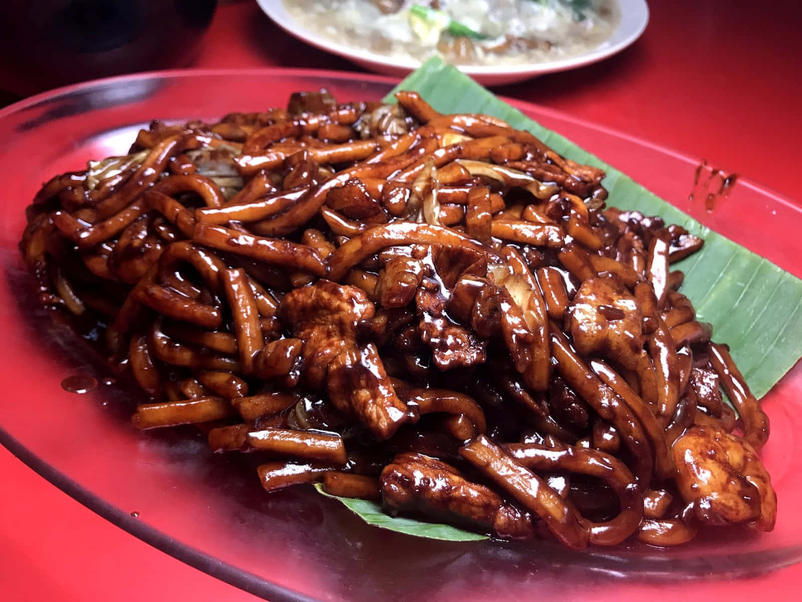 透明盤上的福建麵 壁紙 — 閃亮嘅炒麵配雞肉，濃郁深色醬汁，用紅色碟配香蕉葉上碟。