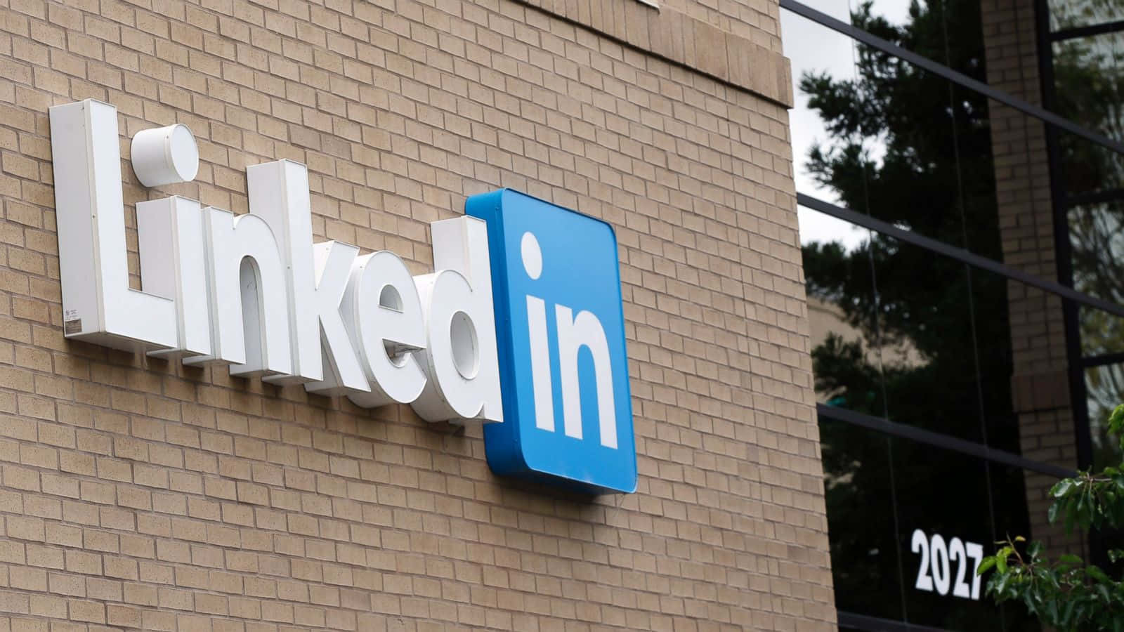 透過 LinkedIn 實現專業成長 壁紙 — 一個大型白色和藍色的LinkedIn標誌安裝在米色磚砌建築上，玻璃窗反射樹木。