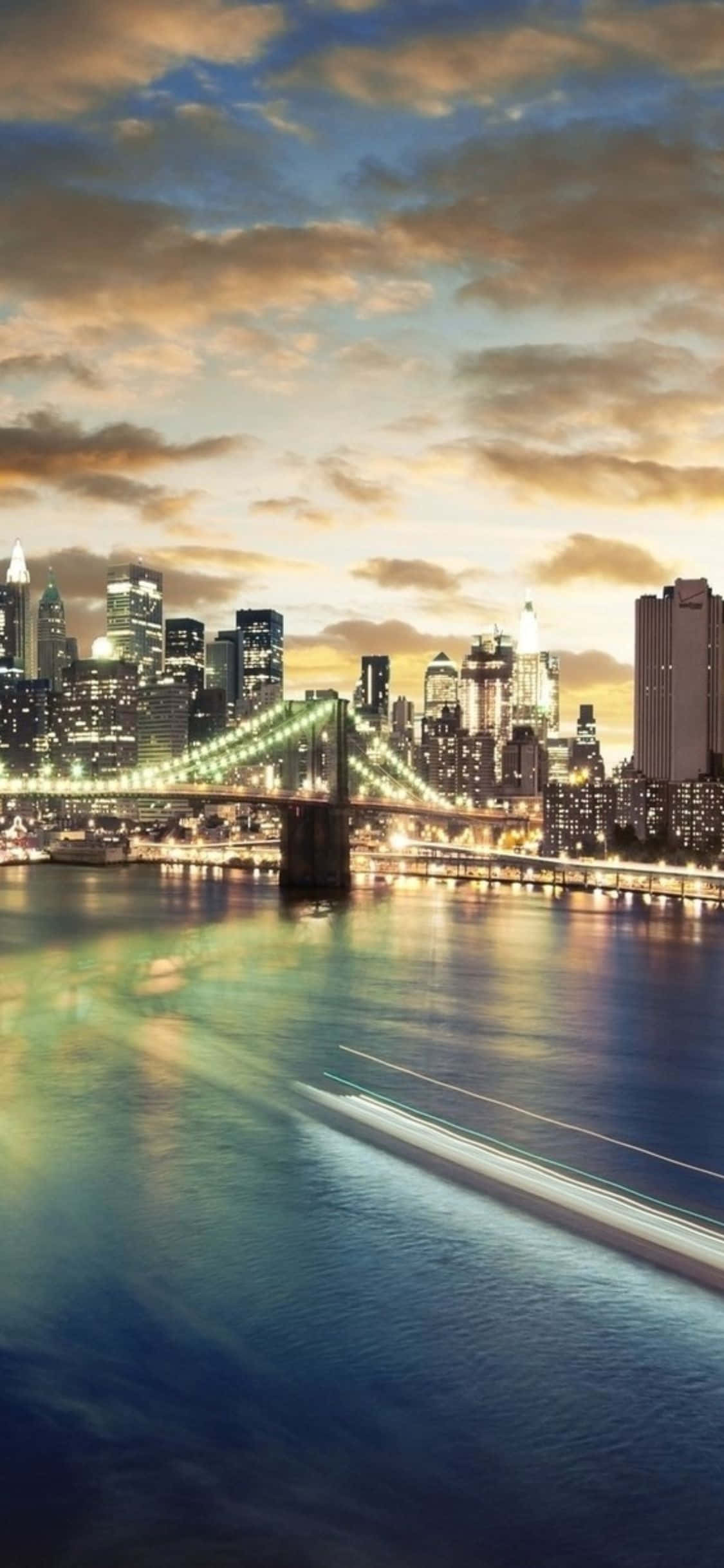 透過iPhone XS鏡頭欣賞紐約市的夜景天際線 壁紙 — 布魯克林大橋和紐約天際線的壯麗黃昏景色，閃爍的城市燈光倒映在下方水面上。