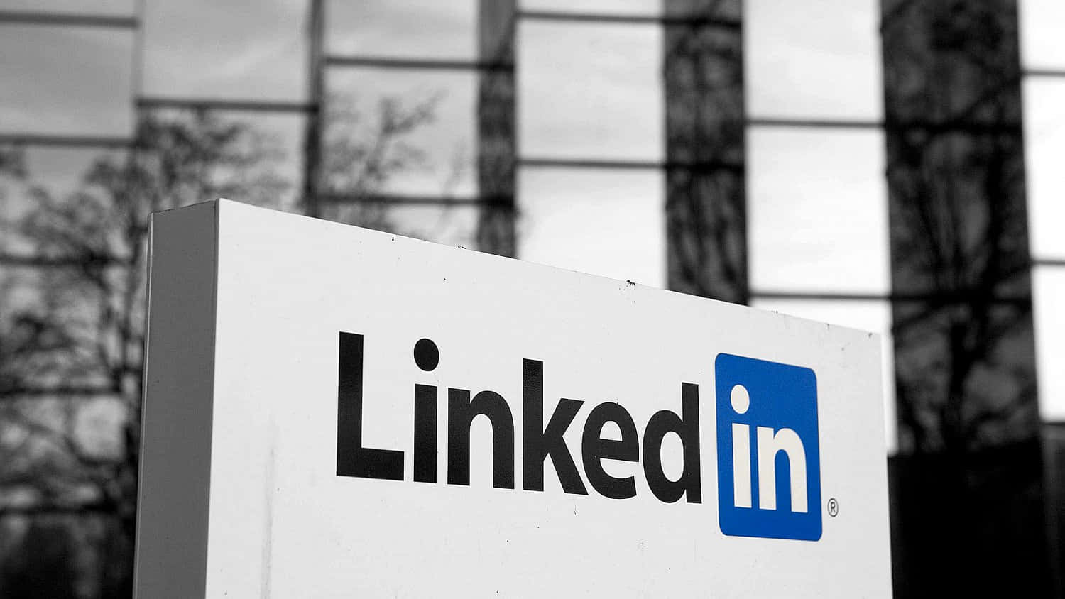透過人脈拓展新機會 壁紙 — 一個黑白相間的LinkedIn標誌矗立在玻璃建築前，映照著光禿禿的樹木倒影。