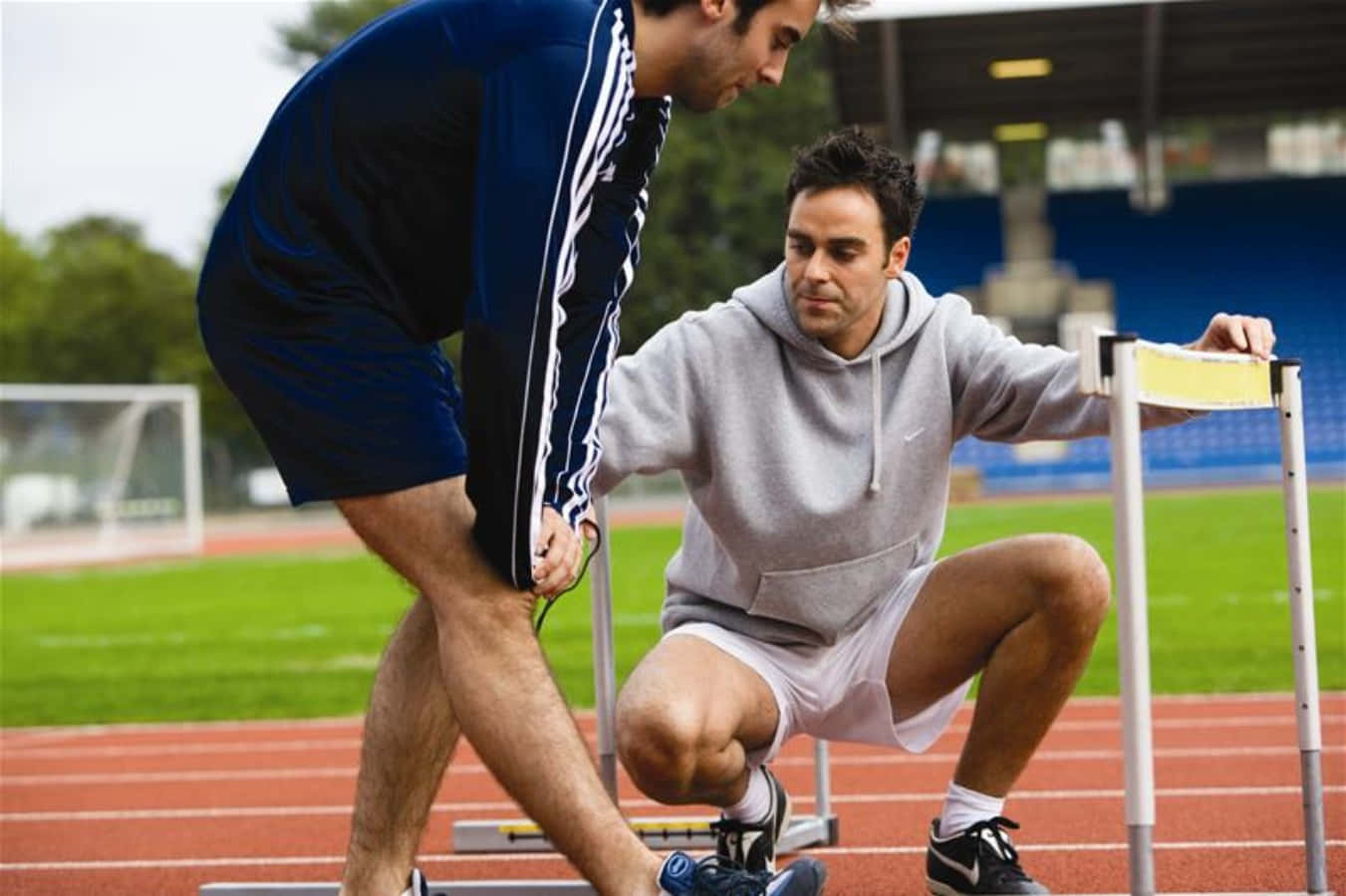 透過個人 Coaching 達成成功 壁紙 — 教練在紅色跑道上調整運動員在跨欄旁的姿勢，背景有綠色場地和體育場座位。