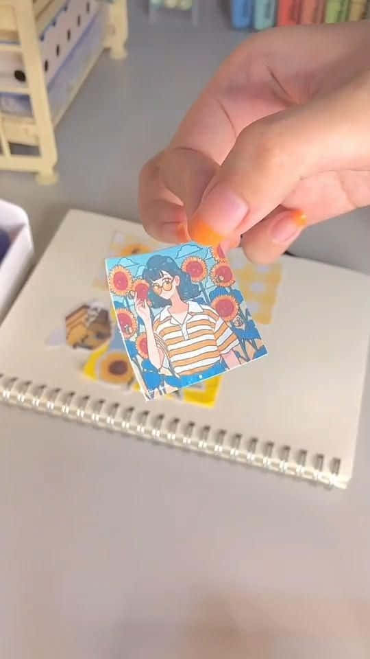 透過剪貼簿表達你的創意 壁紙 — 一隻手在桌上嘅螺旋筆記本上面，拎住一個喺向日葵田嘅女仔嘅彩色貼紙。
