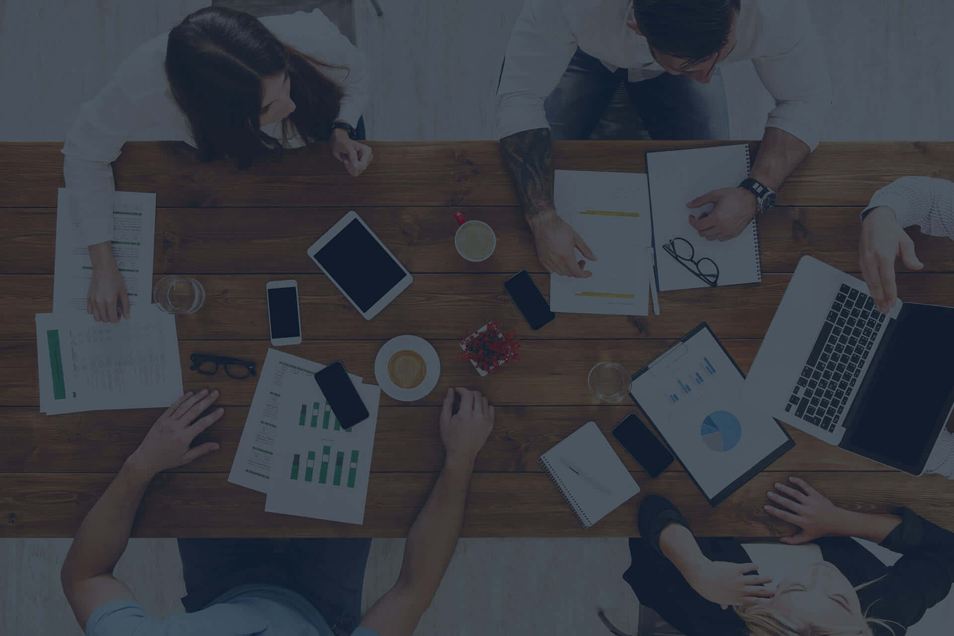 透過努力釋放潛能 壁紙 — Top-down view of four people collaborating around a wooden table filled with laptops papers and coffee cups.