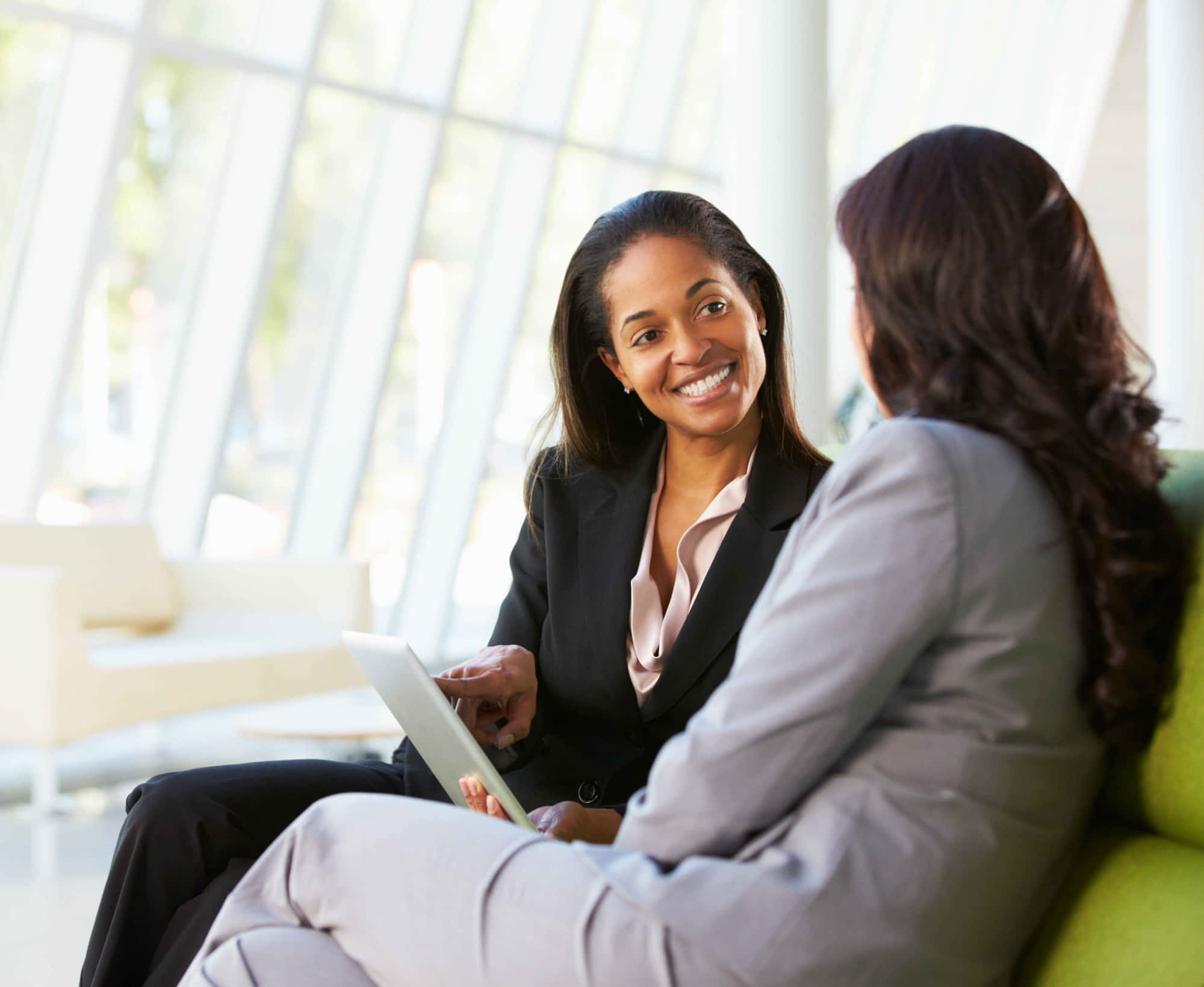 透過專業 Coaching 探索無限可能 壁紙 — 兩位女商人在明亮現代嘅辦公室開會，其中一位拎住平板電腦溫暖咁笑。