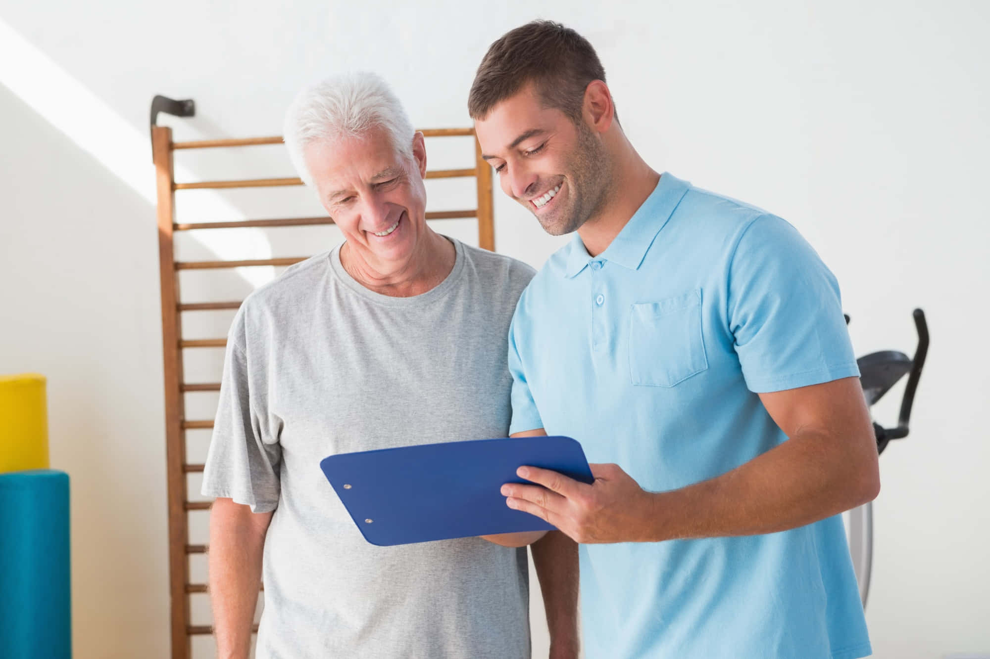 透過專業 Coaching 達成目標 壁紙 — 一位微笑的年輕男子身穿藍色Polo衫，拿著剪貼板，在健身房向一位年長男子展示某樣東西。