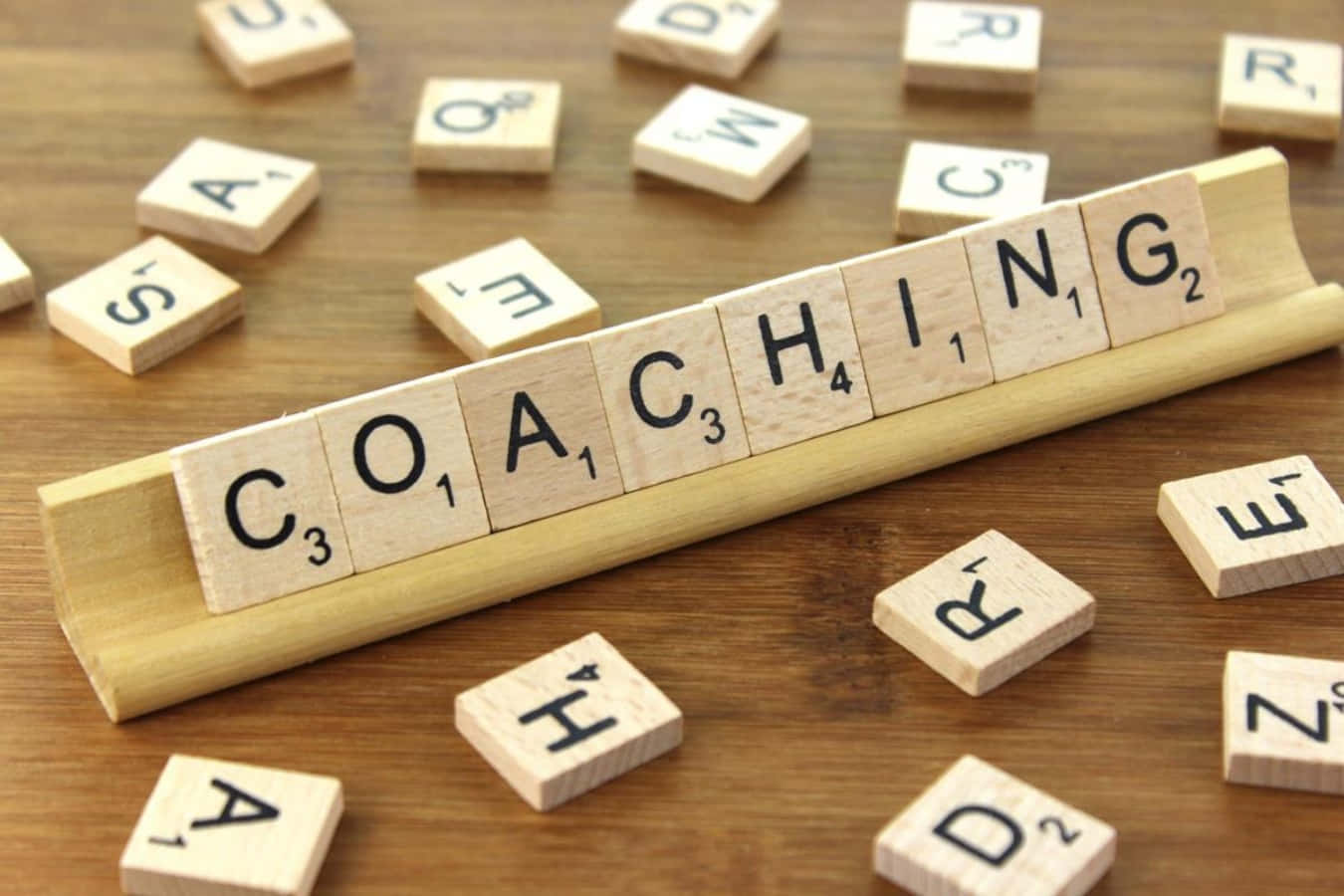 透過生活教練挖掘真正的潛能 壁紙 — 木製 Scrabble 字母牌在木架上拼出 COACHING，置於溫暖的木桌上，周圍散落著字母牌。
