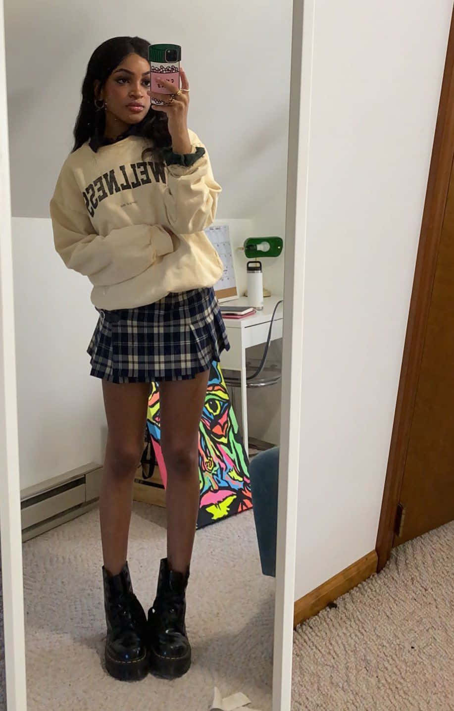 年輕女性穿著奶油色衛衣、格子裙和厚底靴子對著鏡子自拍。