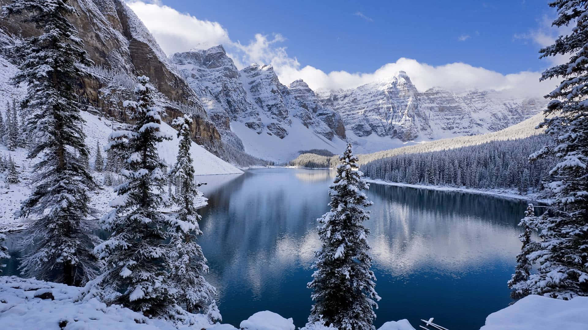 透過這幅壯麗全景景觀，發現冬季的魔力 壁紙 — 一個寧靜的雪山湖泊倒映著高聳的山峰和常綠樹木，在明亮的藍天和蓬鬆的白雲下。