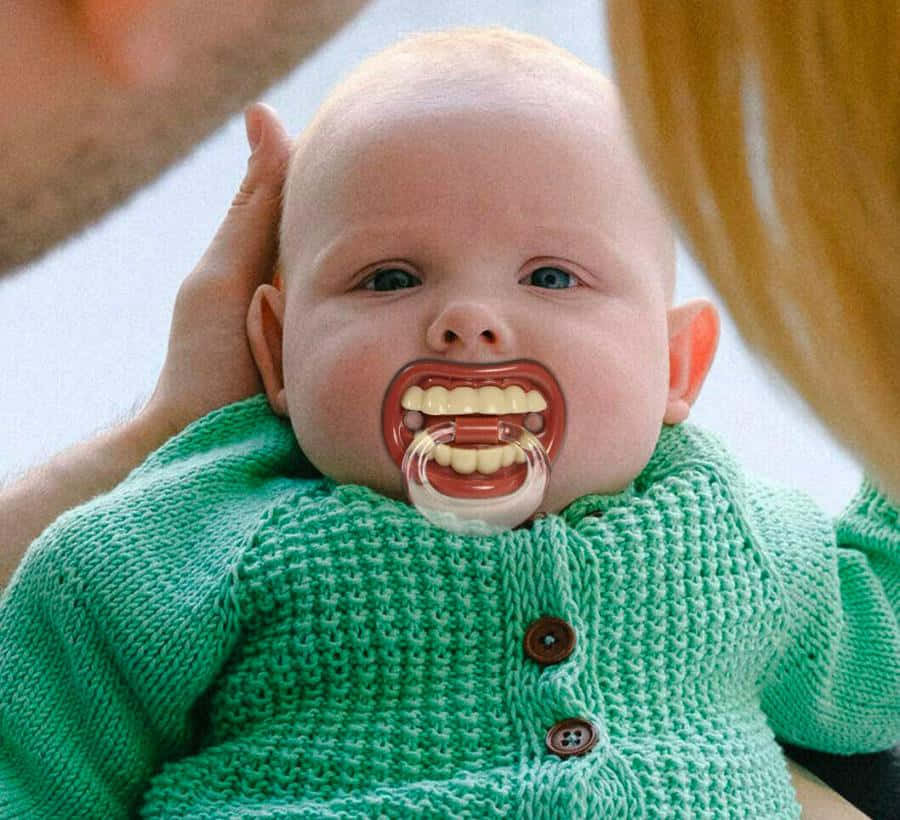 「這太好笑了！」 壁紙 — 一個穿著綠色針織毛衣的嬰兒看著鏡頭，嘴裡含著一個微笑形狀的奶嘴。