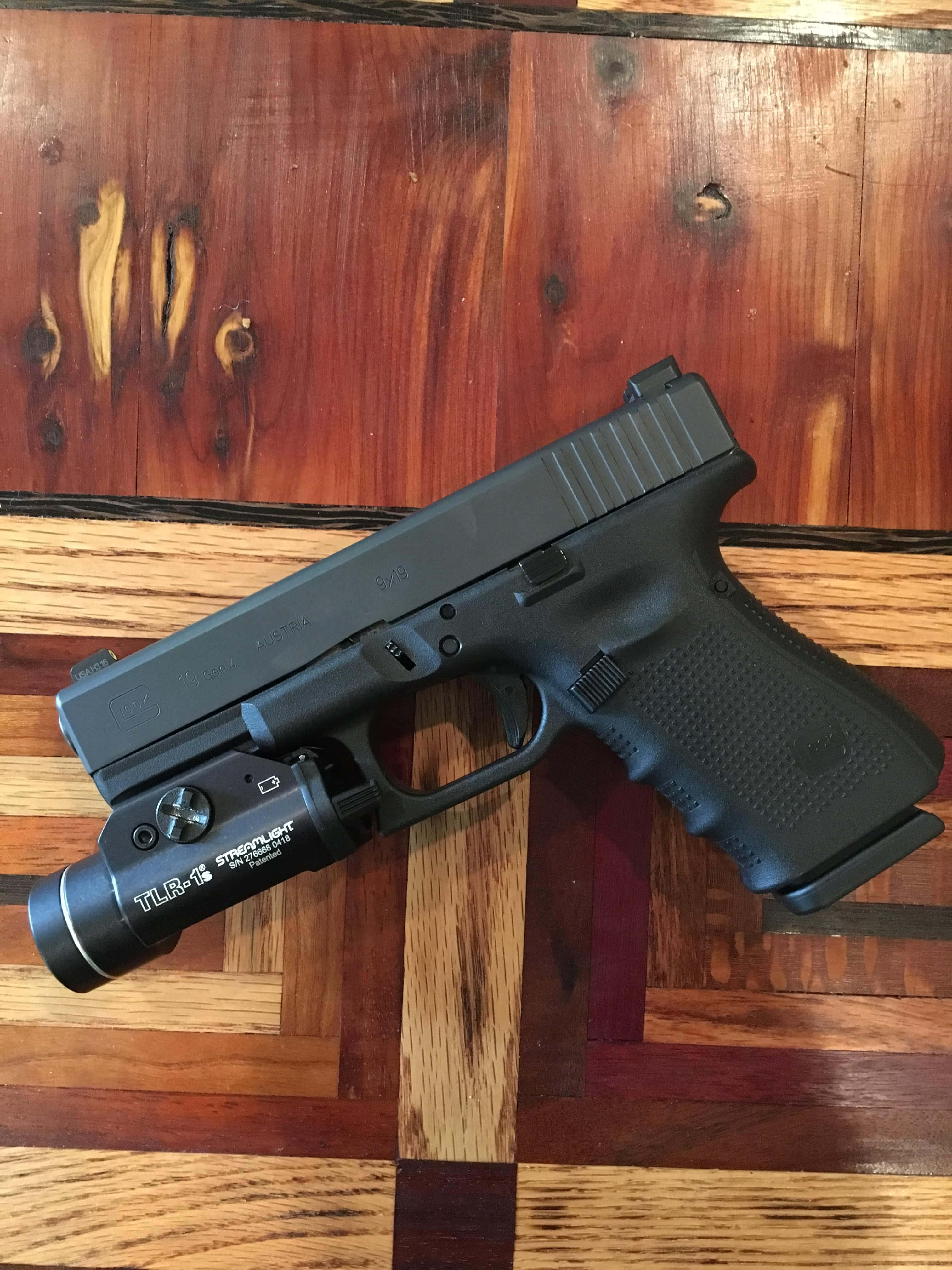 這把 Glock 19 已準備就緒 壁紙 — 一枝黑色手槍連戰術燈放在一塊紋理豐富、帶有天然木節和不同色調的木質表面上。