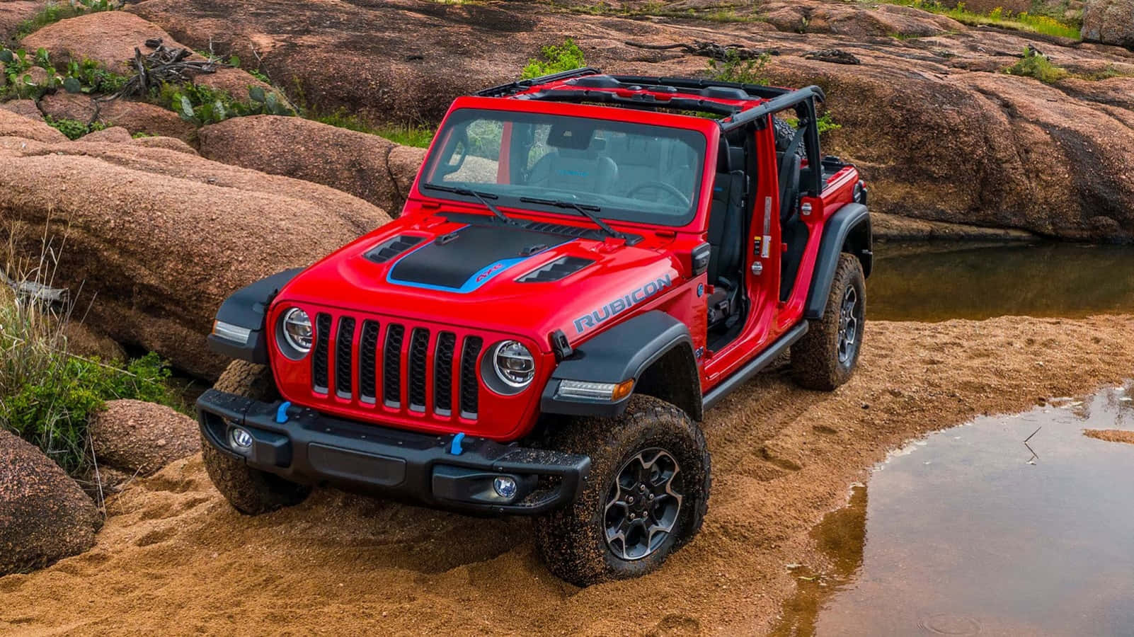 這款堅韌的Jeep Wrangler，隨時準備征服任何地形 壁紙 — 一輛鮮紅色的Jeep Wrangler Rubicon在岩石沙漠溪流中堅固地站立，輪胎沾滿泥濘，車門敞開。