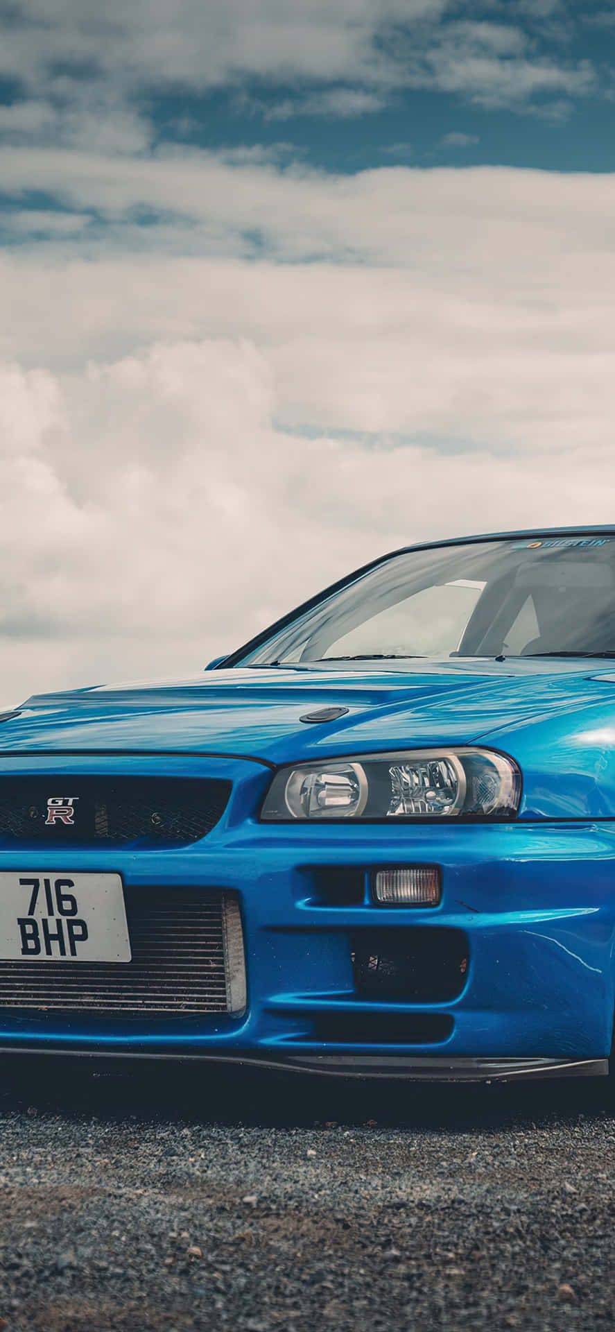藍色日產Skyline R34跑車停放在礫石上，天空多雲。