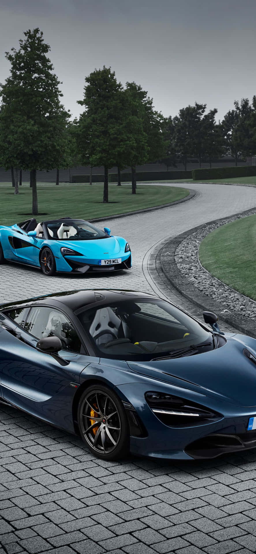 速度與力量的完美結合：iPhone XS Max 與 McLaren 720S 壁紙 — 兩輛時尚的McLaren跑車，一輛藍色開篷車和一輛深藍色轎跑車，停泊在彎曲的石路上，周圍綠樹成蔭，天空多雲。