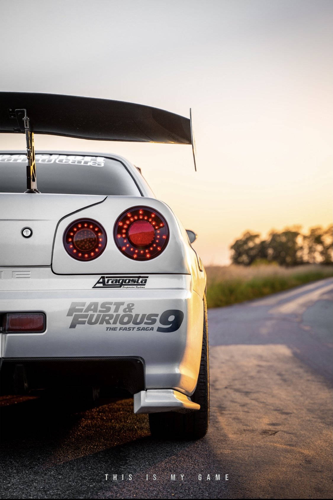 一輛帶有『狂野時速9』貼紙的銀色日產Skyline GT-R，停在日落時分的鄉村道路上。
