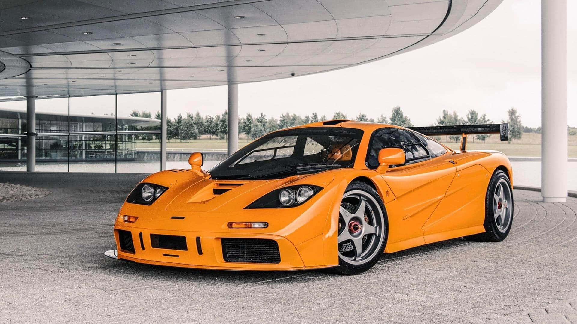 速度與豪華 - 麥拉倫F1 壁紙 — 一輛充滿活力的橙色McLaren F1超級跑車在現代玻璃和鋼製天篷下於日光下閃耀。