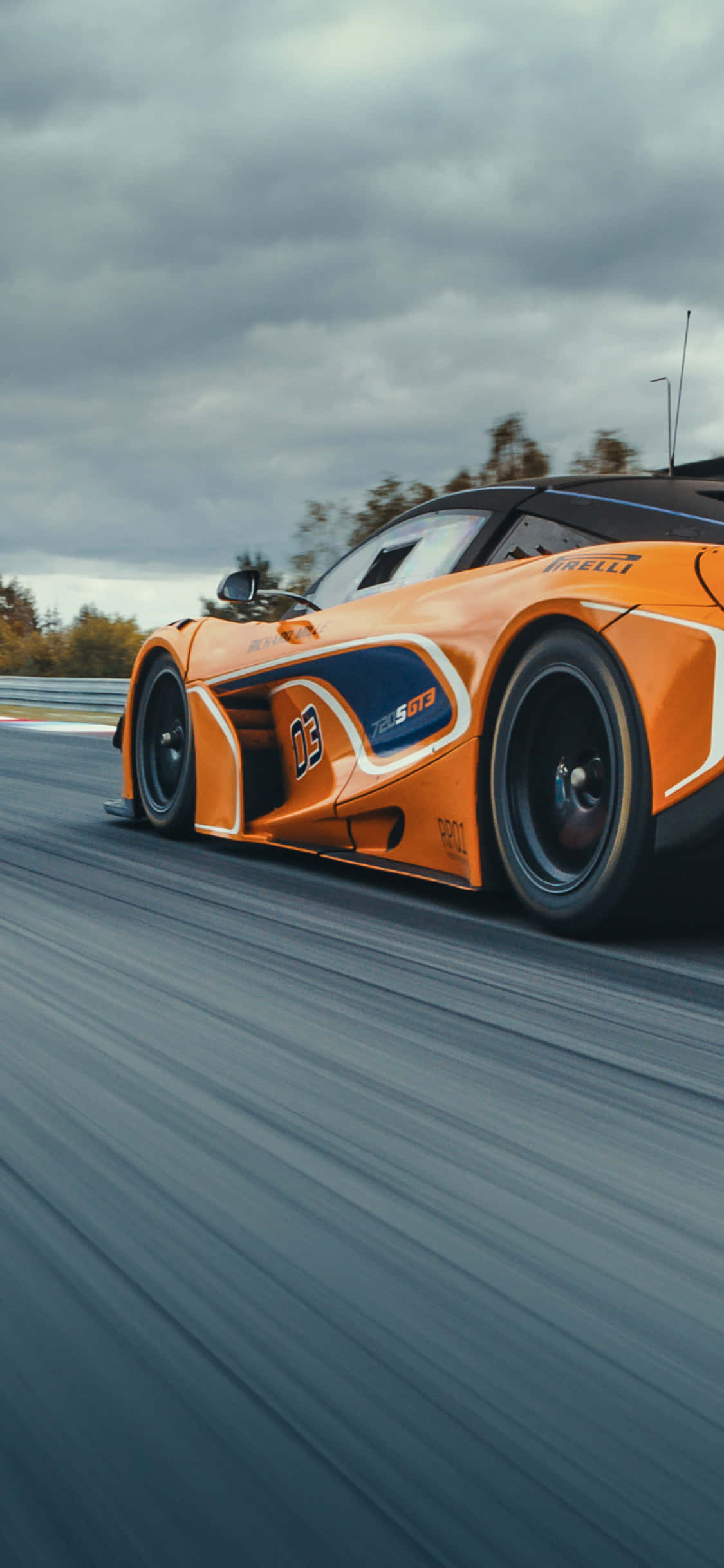 速度與風格——iPhone Xs 與 McLaren 720S 壁紙 — 一輛充滿活力的橙色賽車在潮濕的賽道上奔馳，天空多雲，帶有模糊的動態線條。