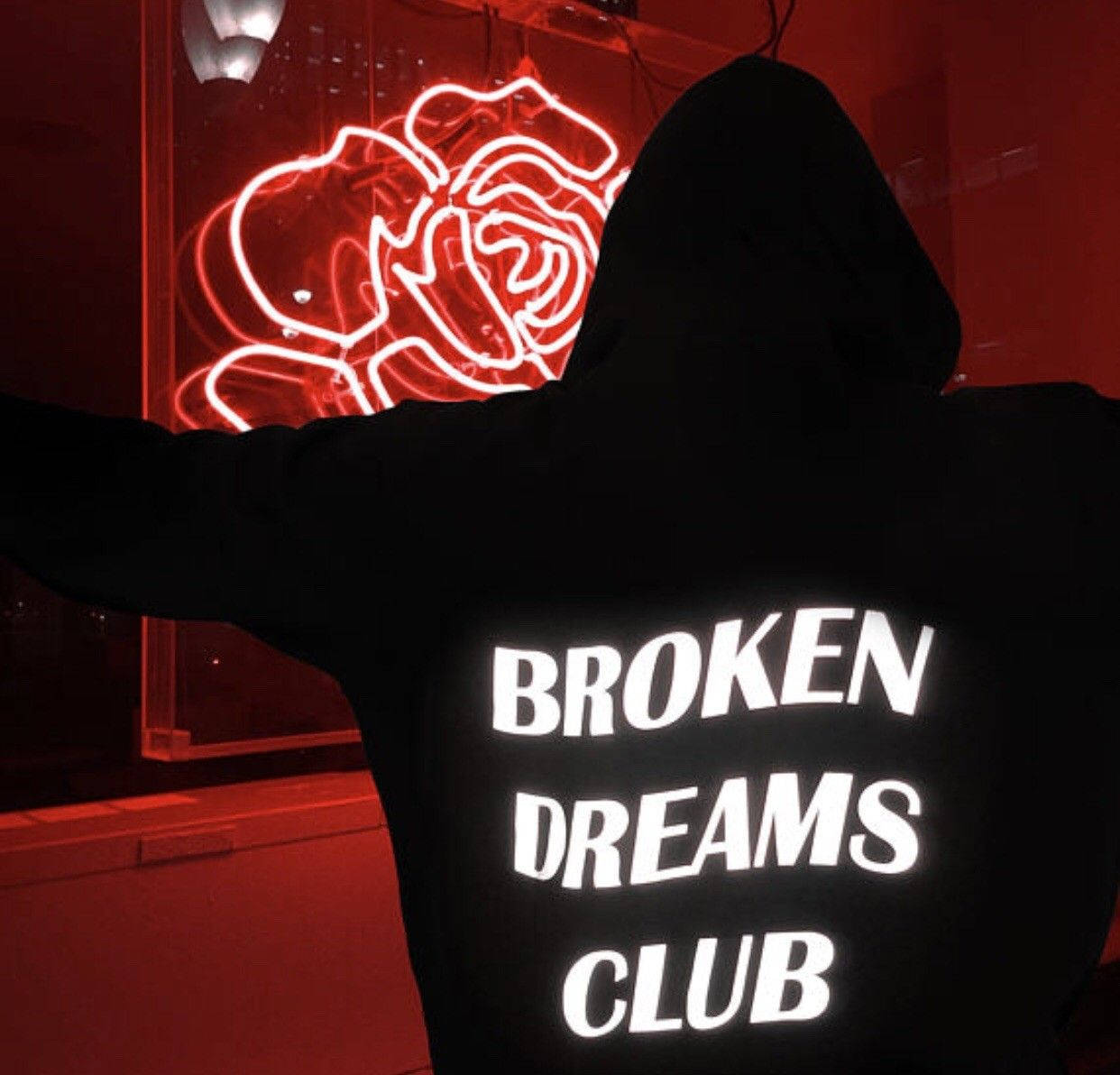 穿著印有「Broken Dreams Club」字樣黑色連帽衫的人，面向紅色霓虹玫瑰。