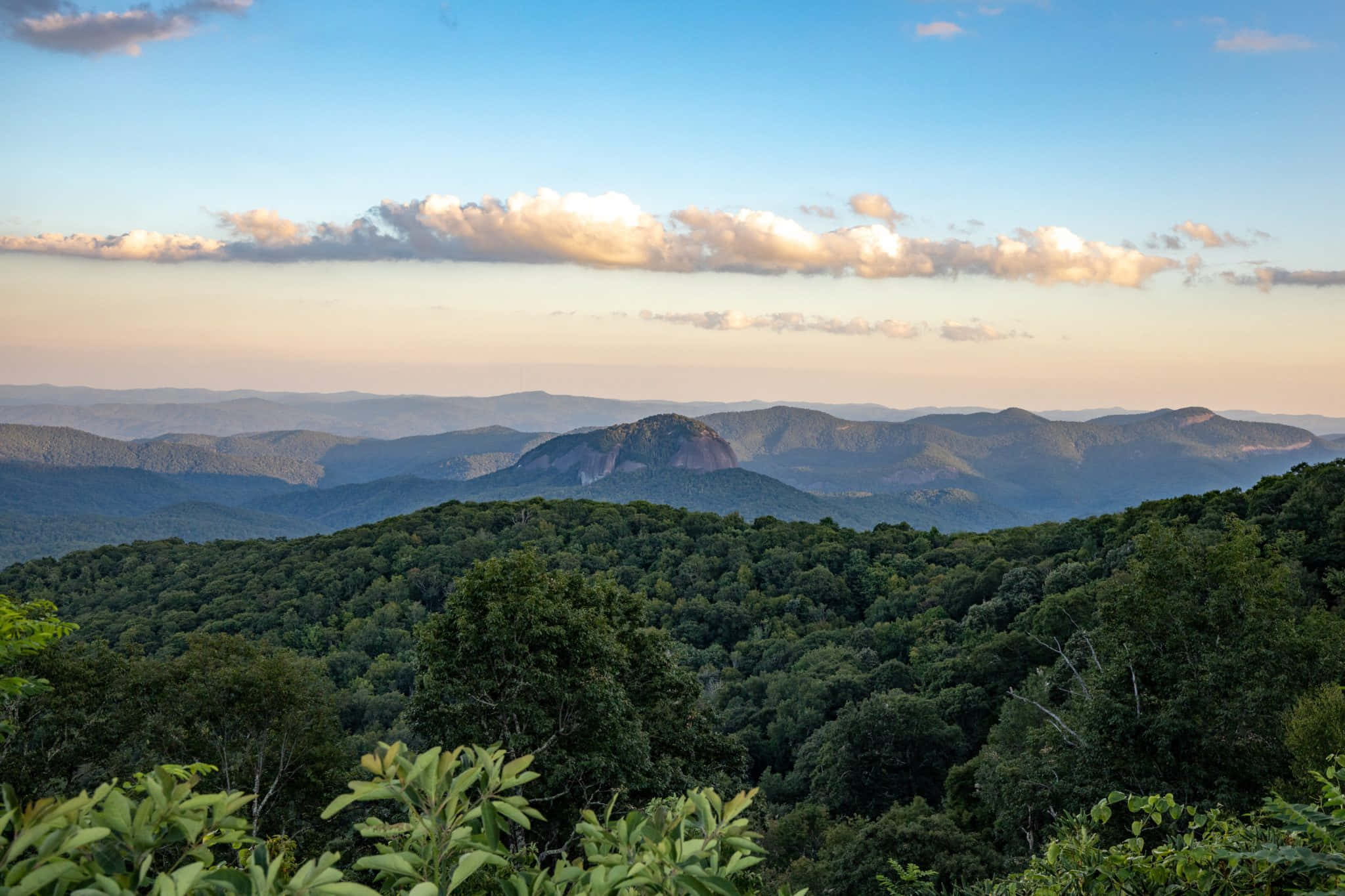 Blue Ridge Mountains 桌布 壁紙 — 連綿起伏的森林山脈在柔和的金色日落天空和零星雲朵下延伸至遠方。