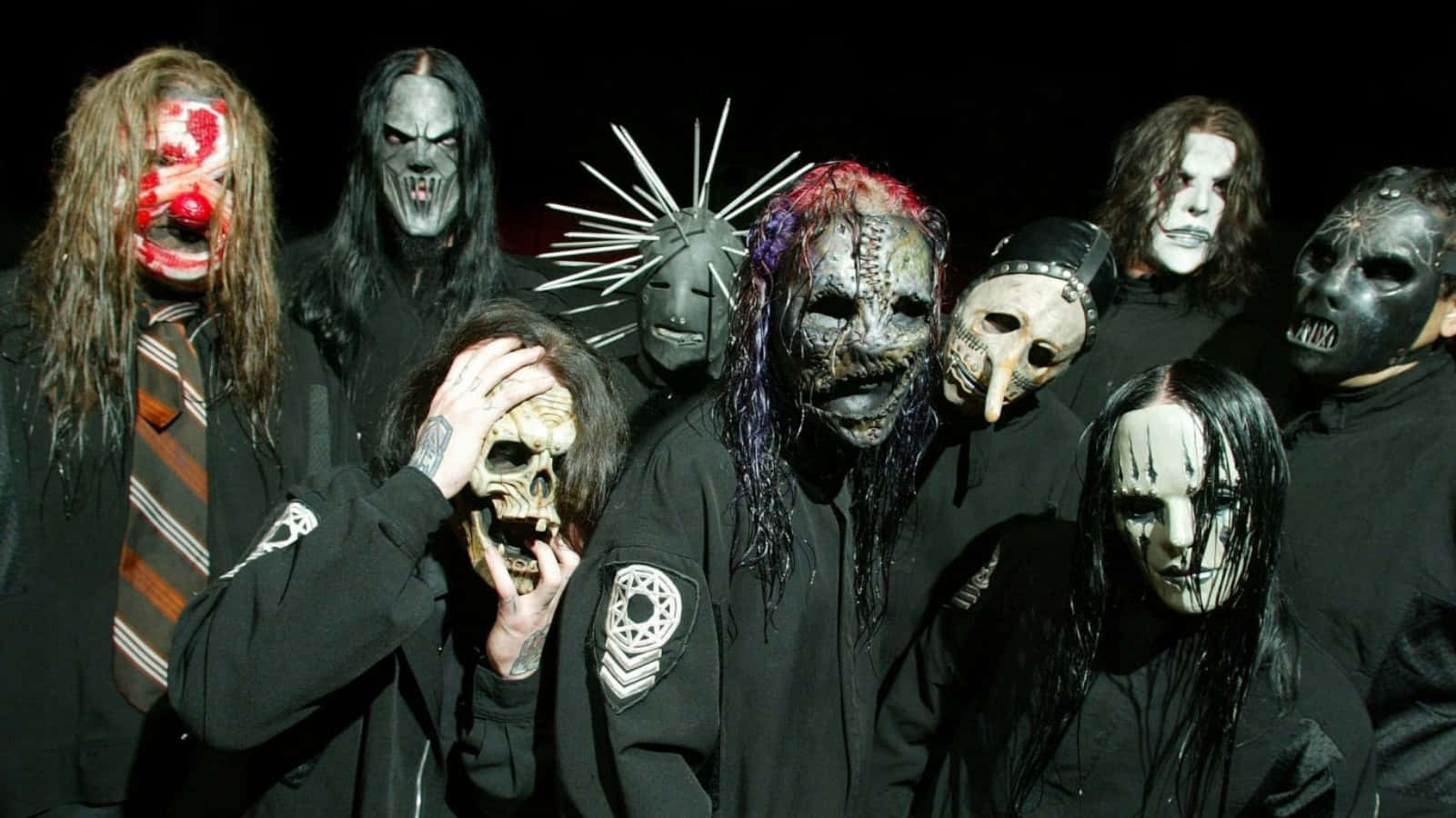 進入Slipknot的世界 壁紙 — 一群戴著怪誕面具、身穿深色衣服的音樂家，在黑色背景下擺出威脅姿勢。