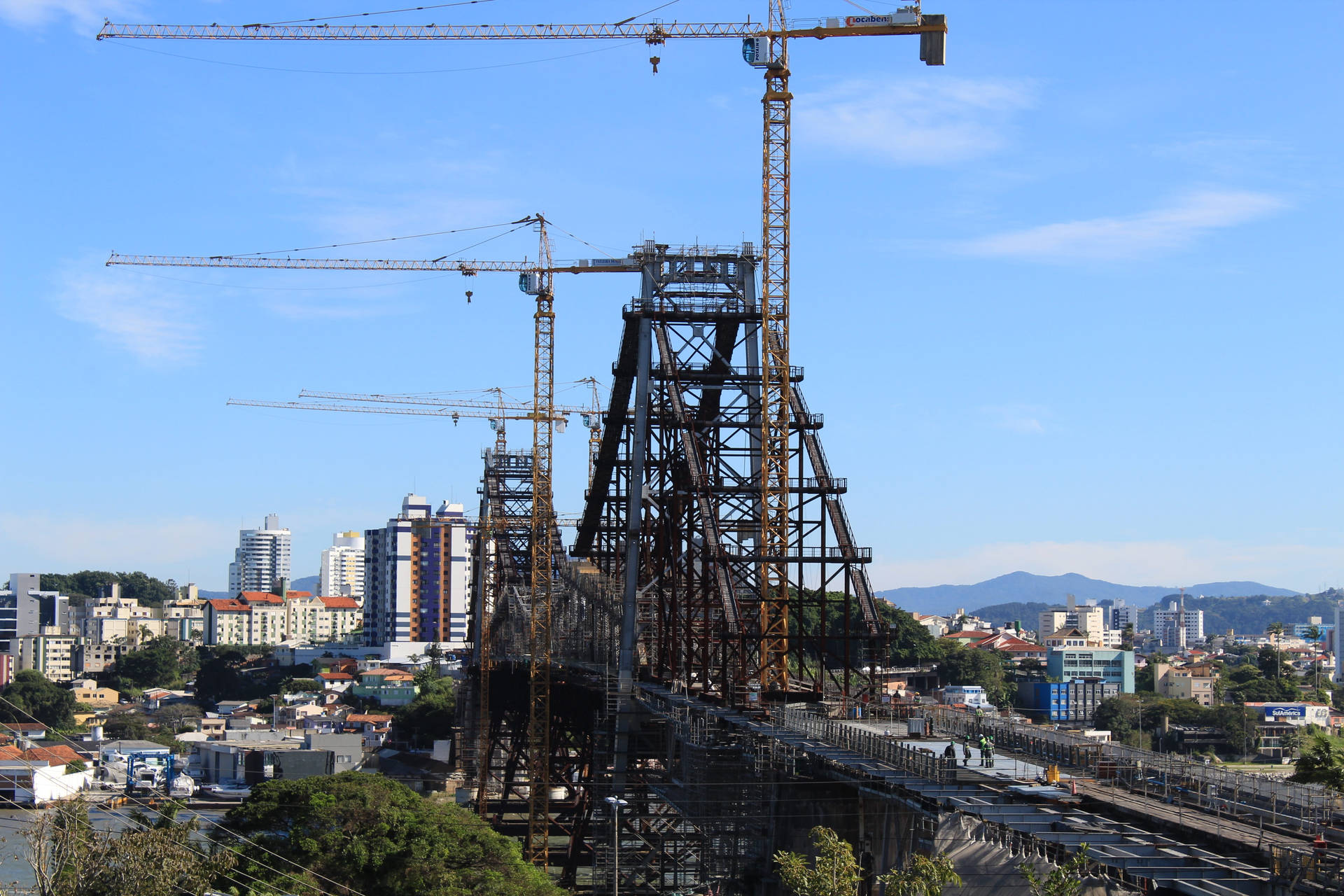 鋼橋正在建造中，起重機在藍天下，城市中。