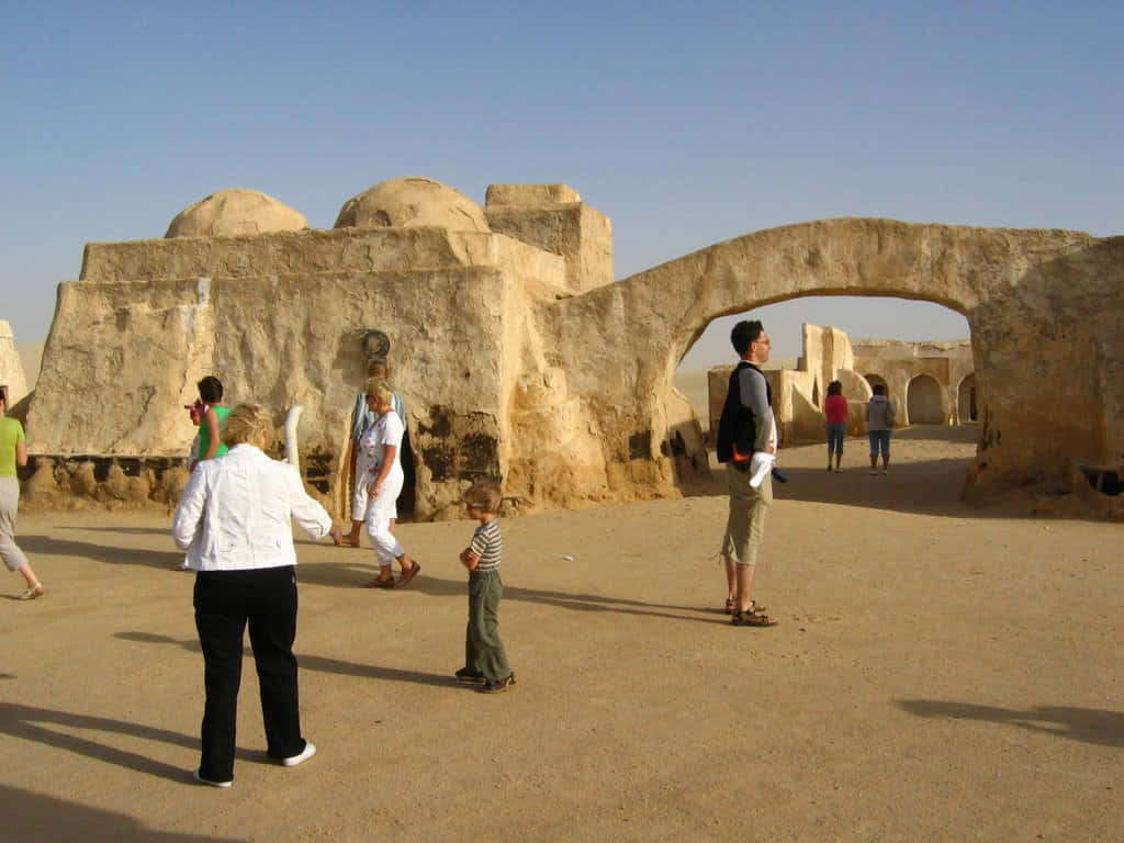 Mos Eisley 桌布 壁紙 — 遊客在晴朗的藍天下探索帶有圓頂結構和石拱門的沙漠遺跡。
