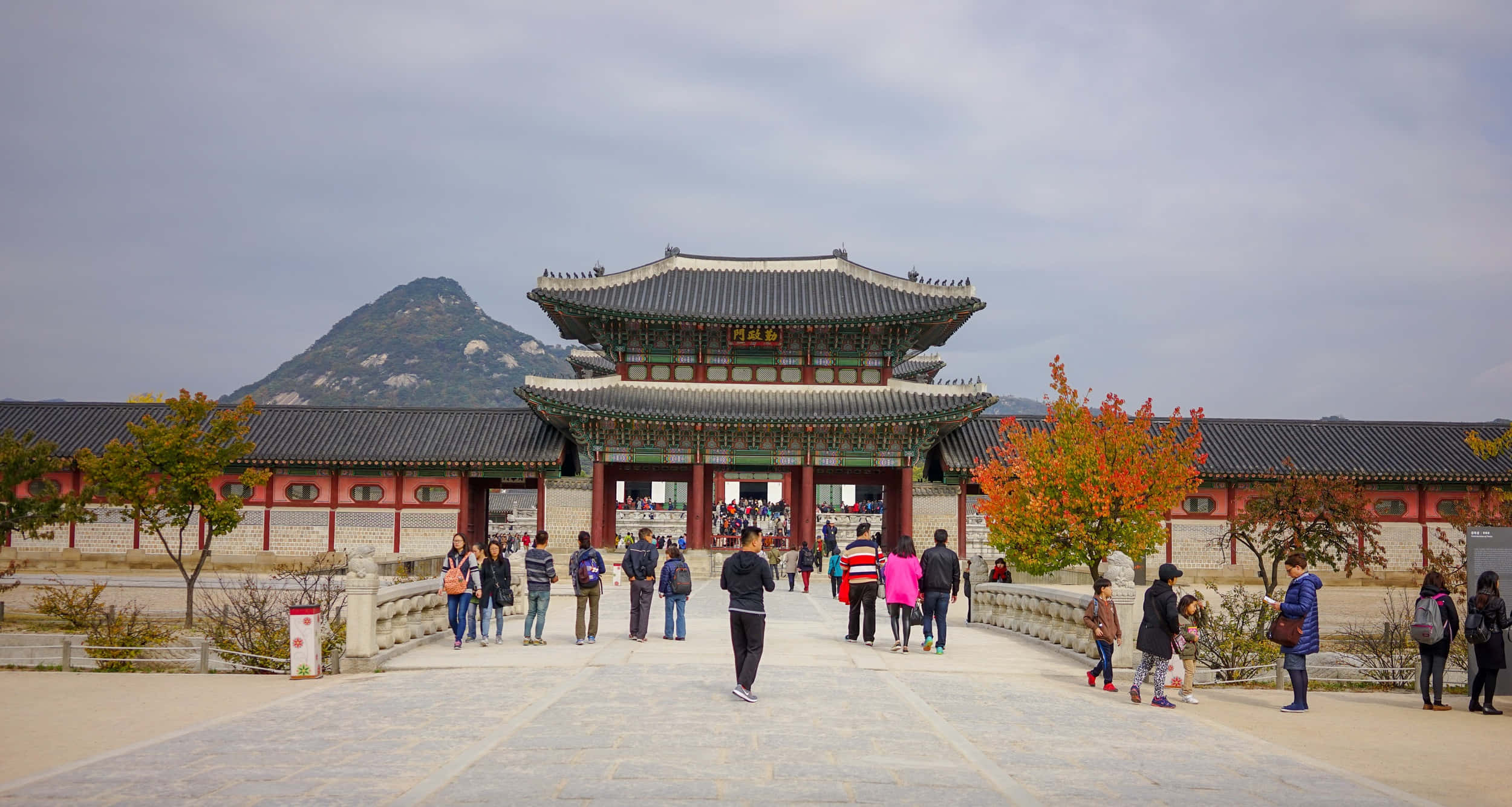 South Korea 桌布 壁紙 — 遊客漫步走過石橋，前往一座傳統韓國宮殿大門，大門有紅色柱子和灰色瓦片屋頂，天空多雲。