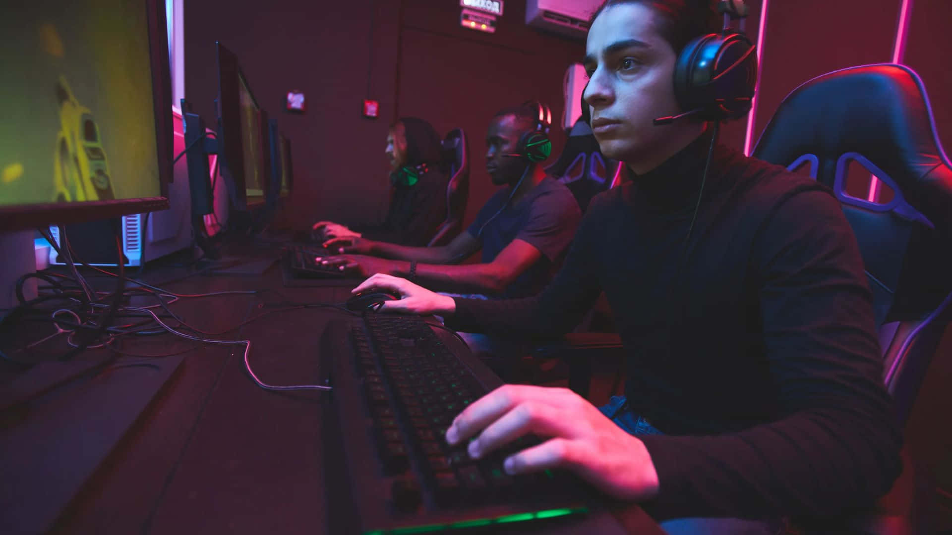 Gaming Tournaments 桌布 壁紙 — 遊戲玩家在昏暗的房間中，被霓虹粉紅和綠色燈光包圍，專注於發光的屏幕。