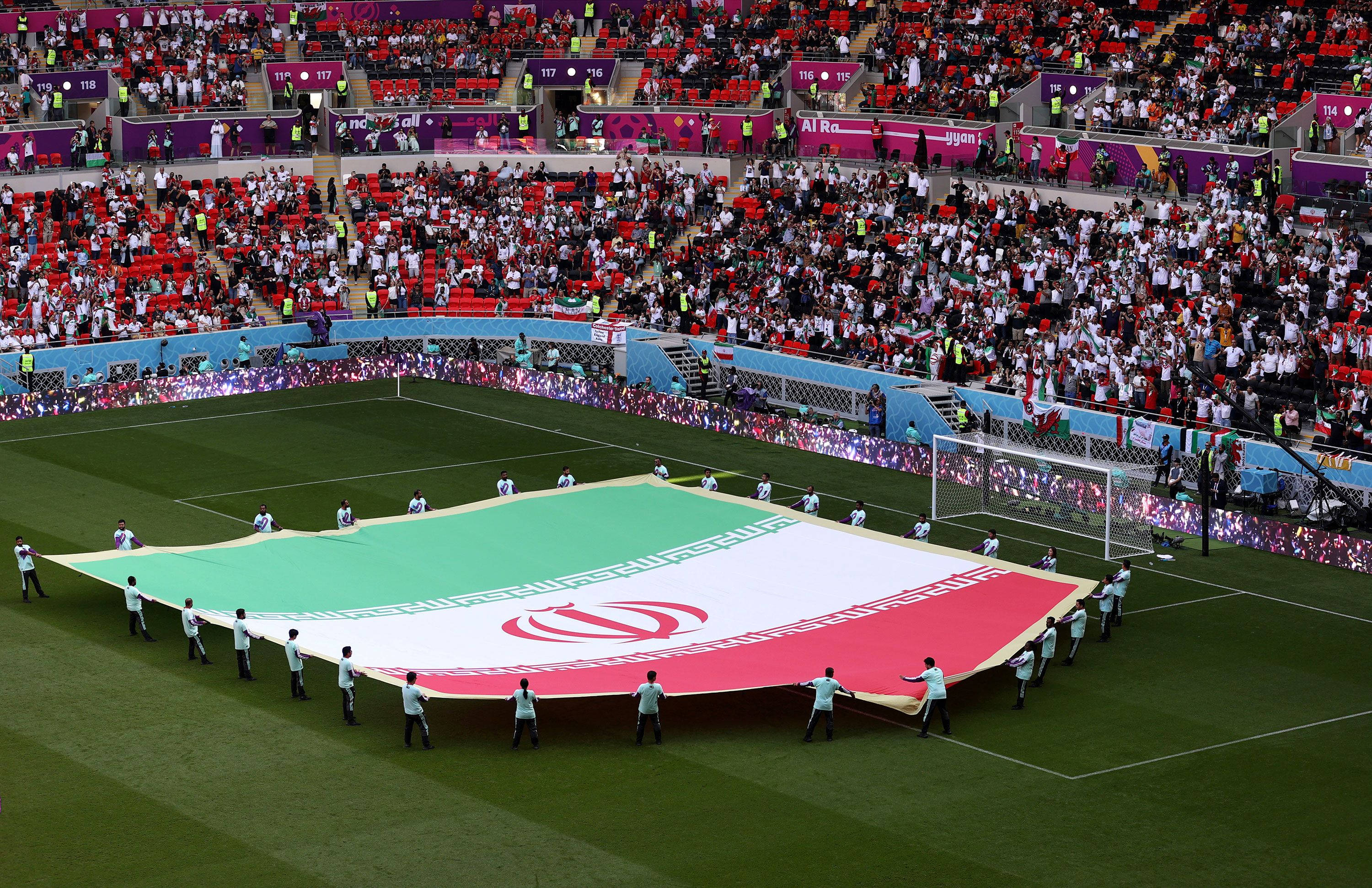 Iran 桌布 壁紙 — 運動員在擠滿觀眾的體育館中，攜帶一面巨大的伊朗國旗穿過充滿活力的綠色足球場。