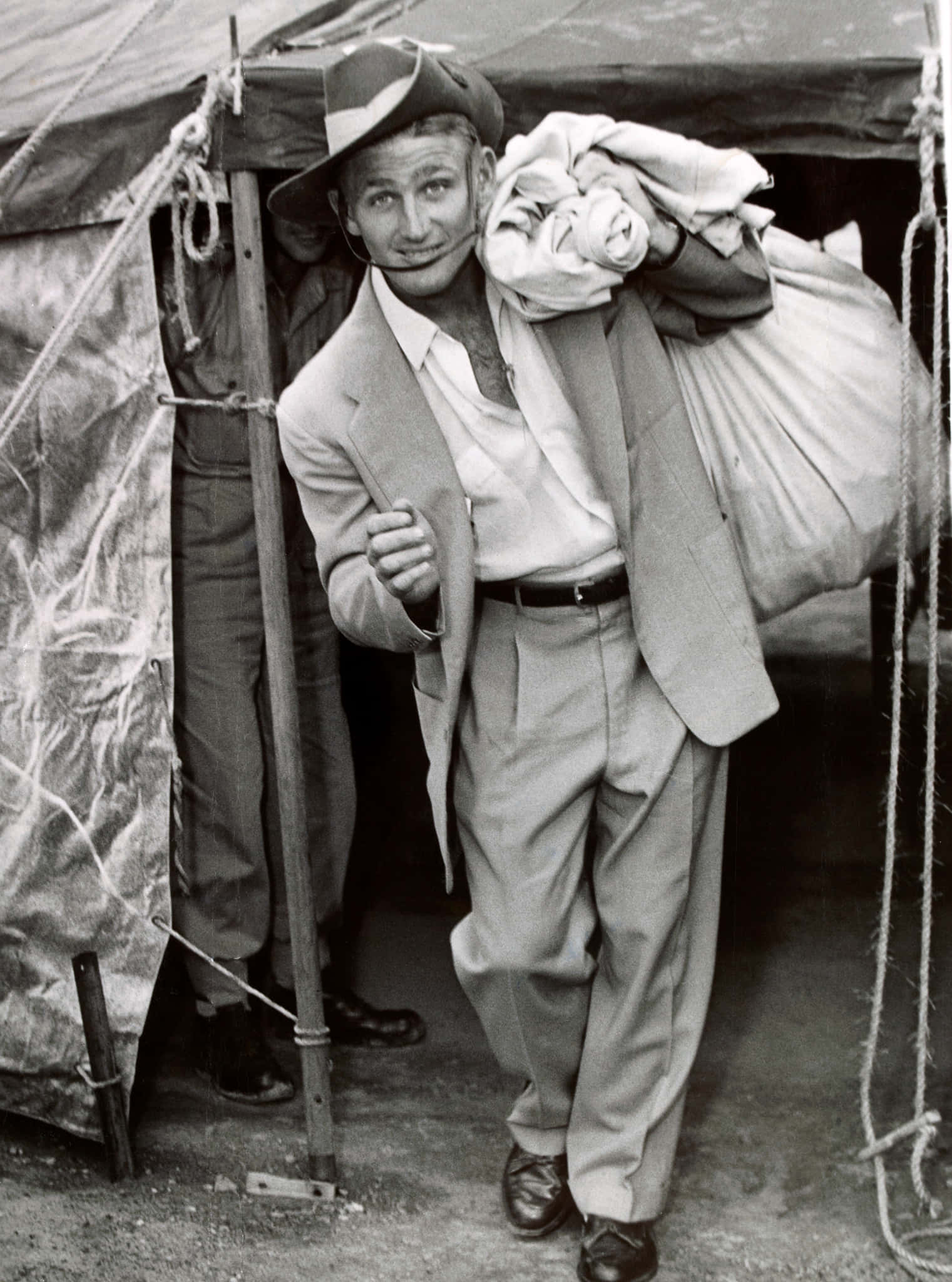 運動員萊·霍德1954年國防服役 壁紙 — 一個戴帽穿西裝的微笑男人，站在帳篷前，抱著一大捆衣服。