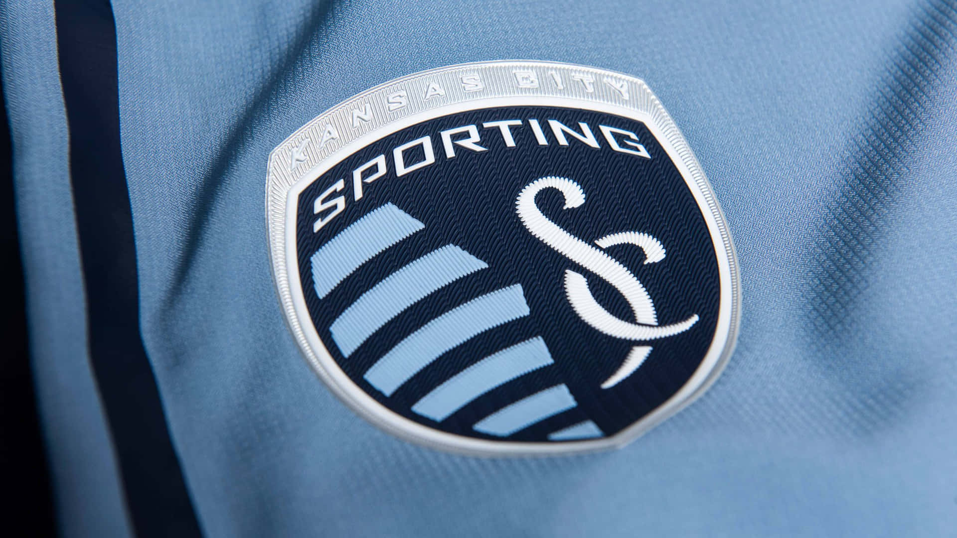 運動家堪薩斯城隊標誌印於球衣上 壁紙 — Sporting Kansas City 足球隊徽章近距離特寫，繡於淺藍色運動球衣布料上。