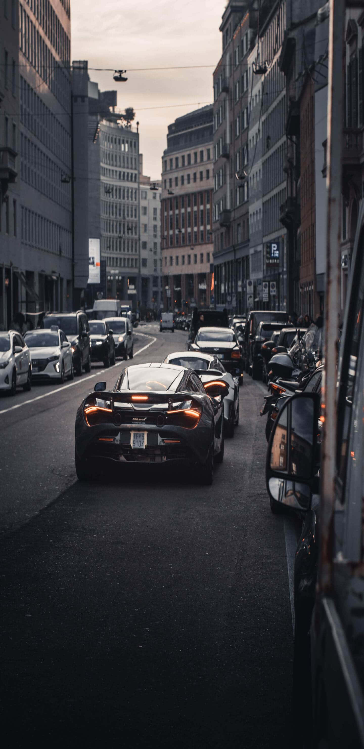 運動車像素 3xl 麥拉倫 720s 城市背景 壁紙 — 一輛時尚的黑色跑車在狹窄的城市街道上行駛，兩旁是高樓大廈和停泊的車輛，天空略顯陰沉。