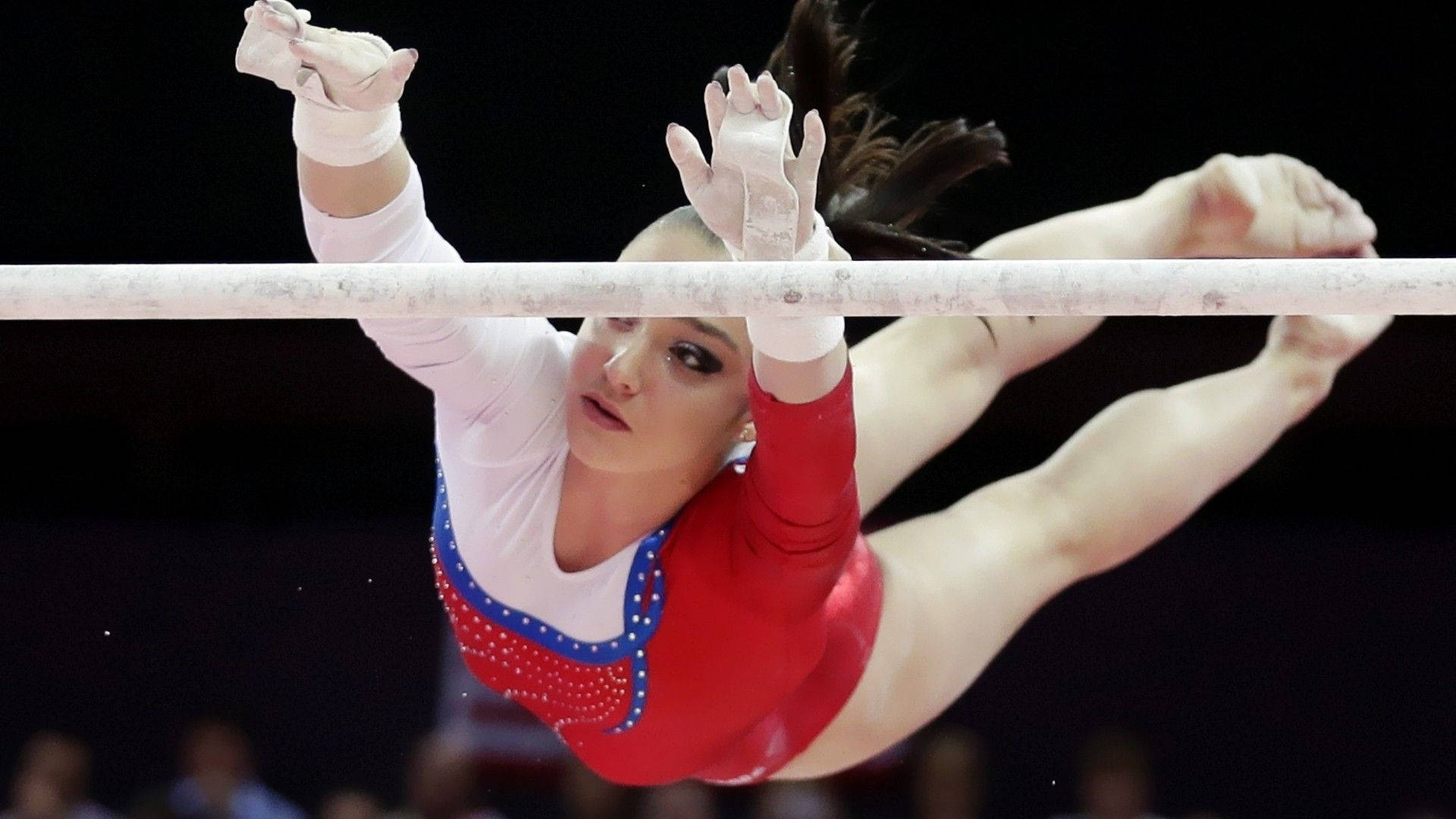 運動體操選手阿莉婭·穆斯塔菲娜 壁紙 — 一名身穿紅白緊身衣的女子體操運動員在黑暗的競技場背景下，專注地抓住高槓擺動中。