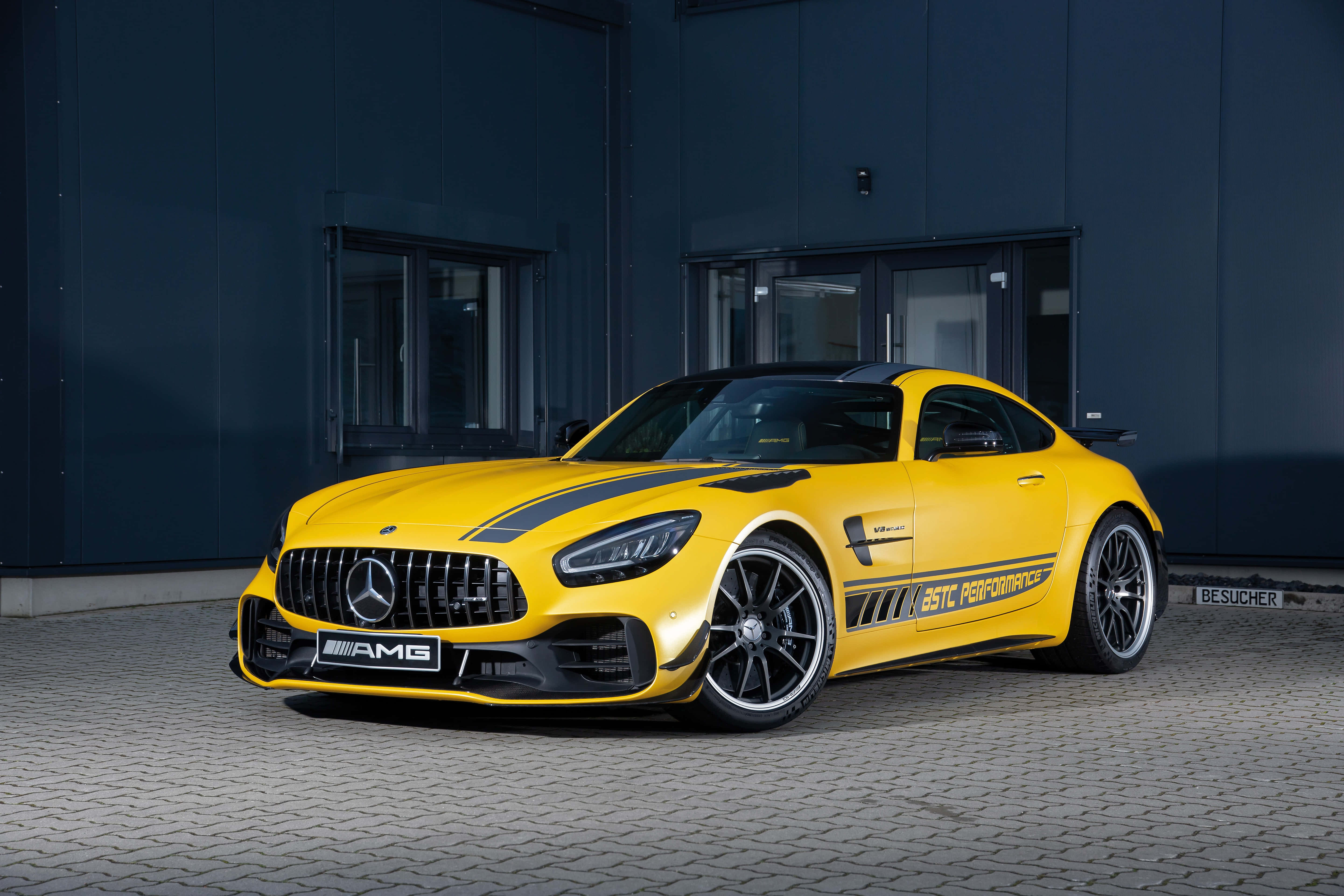 運動黃色梅賽德斯-AMG GT R 車款背景 壁紙 — 一輛鮮黃色AMG GT R跑車帶有黑色賽車條紋，停泊在鋪砌表面上，背景是深藍色工業建築。