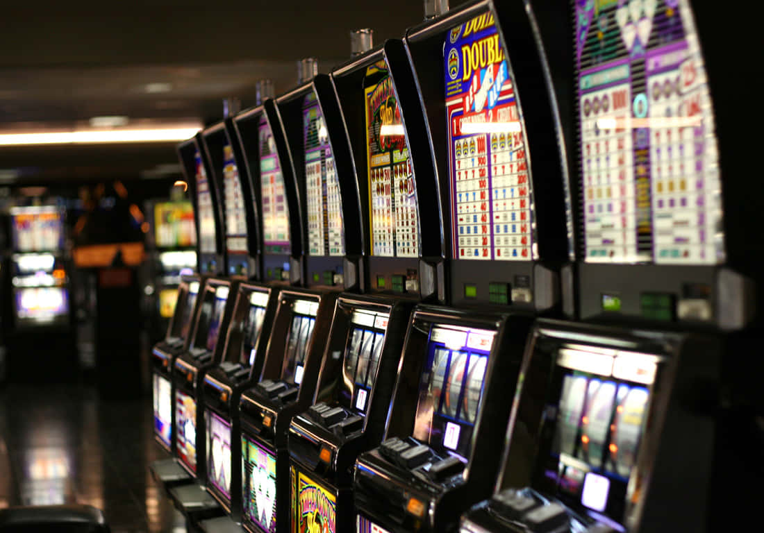 運氣遊戲：令人興奮的插槽機圖像 壁紙 — 一排明亮照明的老虎機，位於燈光昏暗的賭場中，帶有發光的屏幕和色彩繽紛的顯示屏。