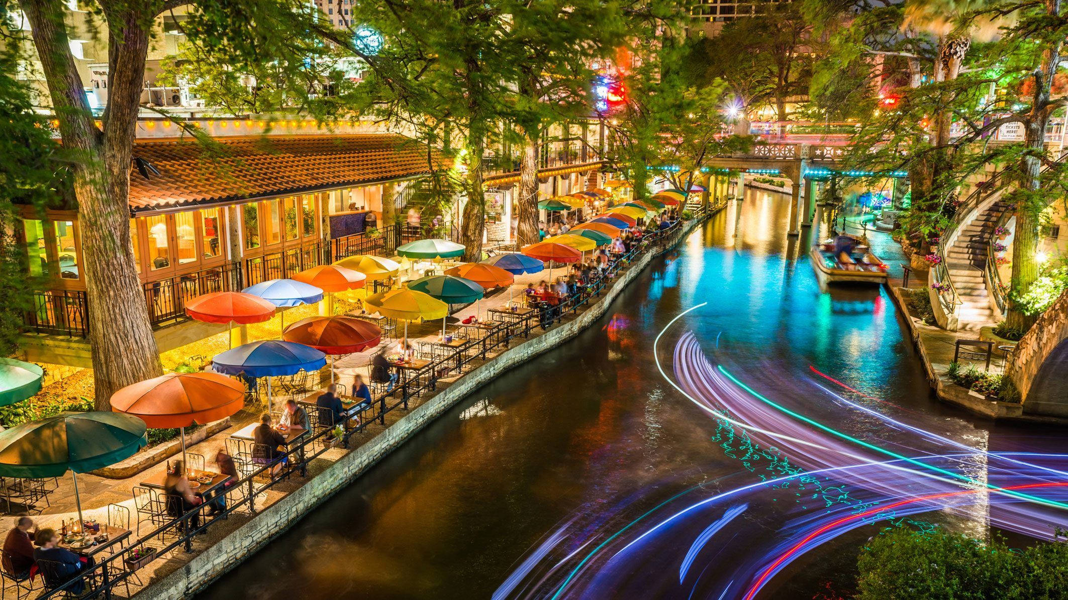 水 桌布 壁紙 — 運河沿岸充滿活力的夜景，色彩繽紛的雨傘在燈串下閃爍，並有船隻經過留下的霓虹燈條紋。