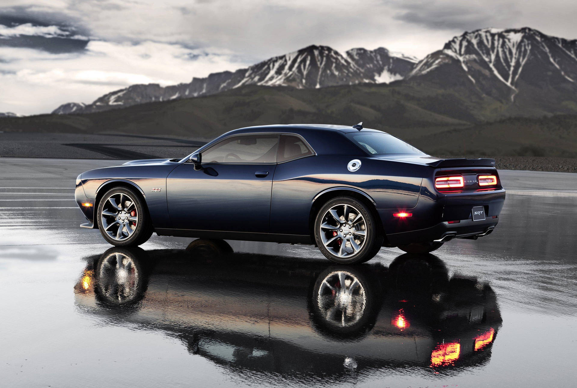 道奇挑戰者 SRT Hellcat 壁紙 — 一輛時尚的深藍色肌肉車在潮濕的路面上反射，背景是雲天下積雪覆蓋的山脈。