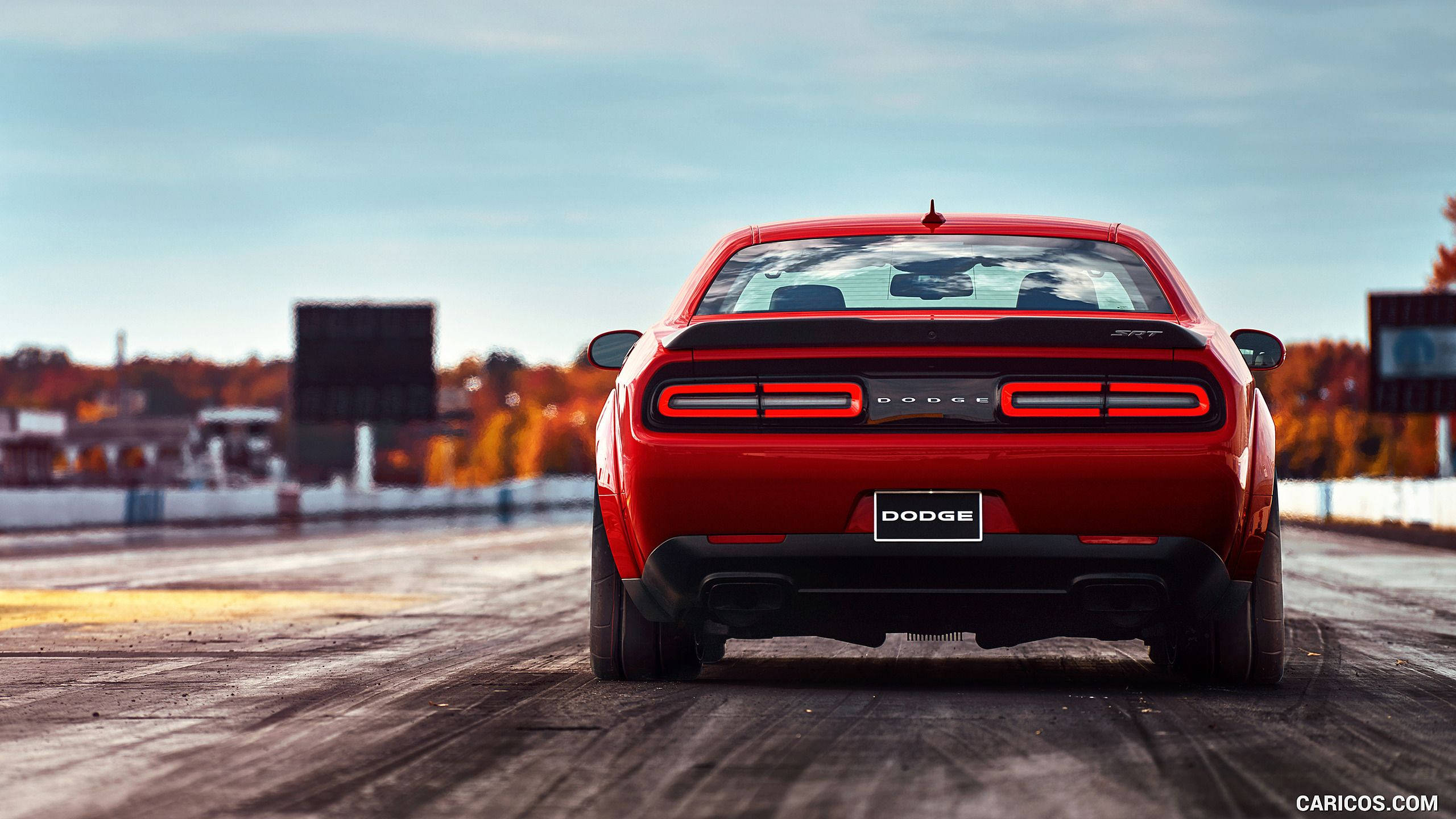 道奇挑戰者於賽道上 壁紙 — 一輛鮮紅色Dodge Challenger SRT Hellcat在賽道上，背景有秋樹和晴朗天空的後視圖。