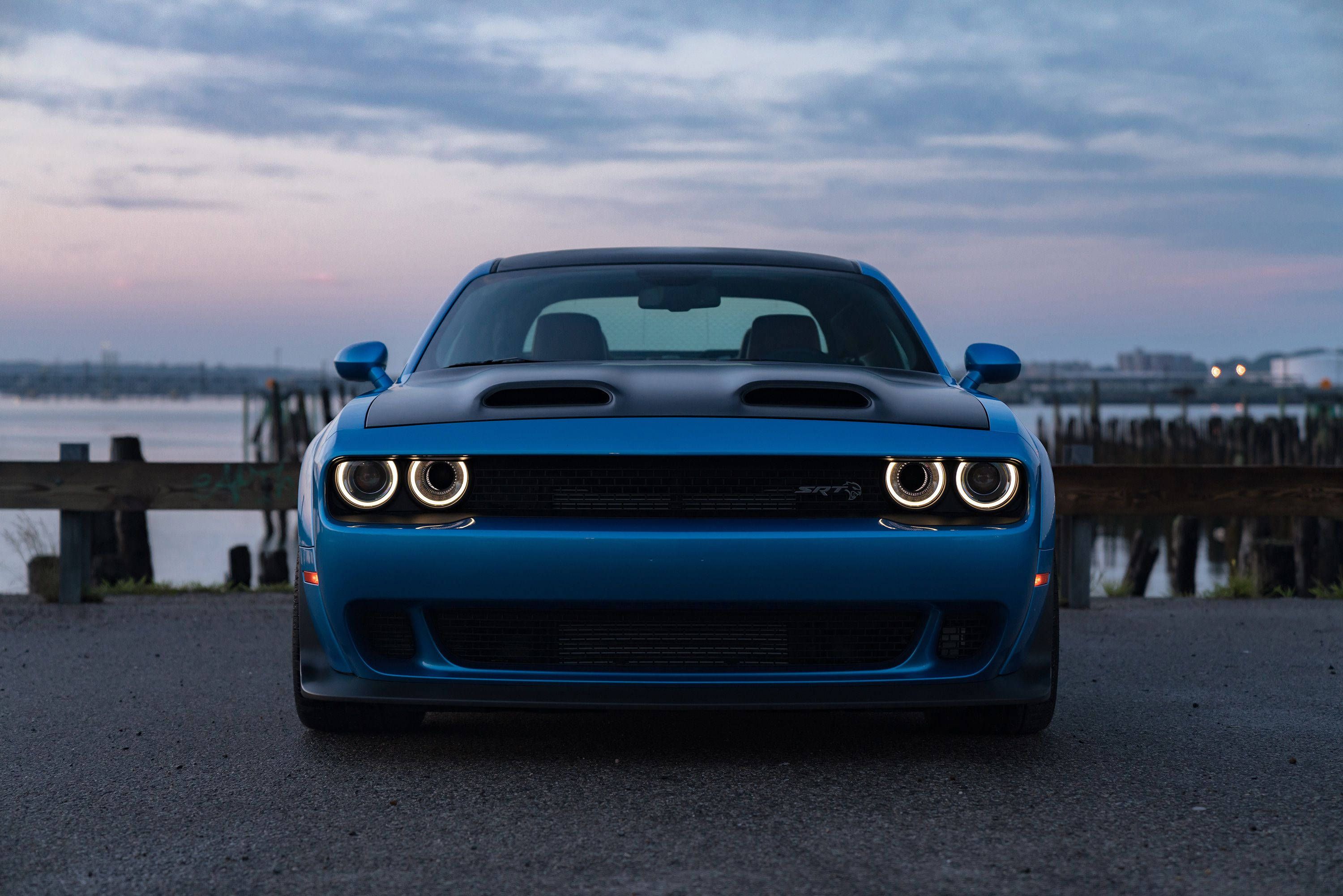道奇挑戰者藍色SRT Hellcat 壁紙 — 一輛充滿活力的藍色道奇挑戰者SRT Hellcat正面朝前，圓形車頭燈發光，背景是暮色中的海濱。