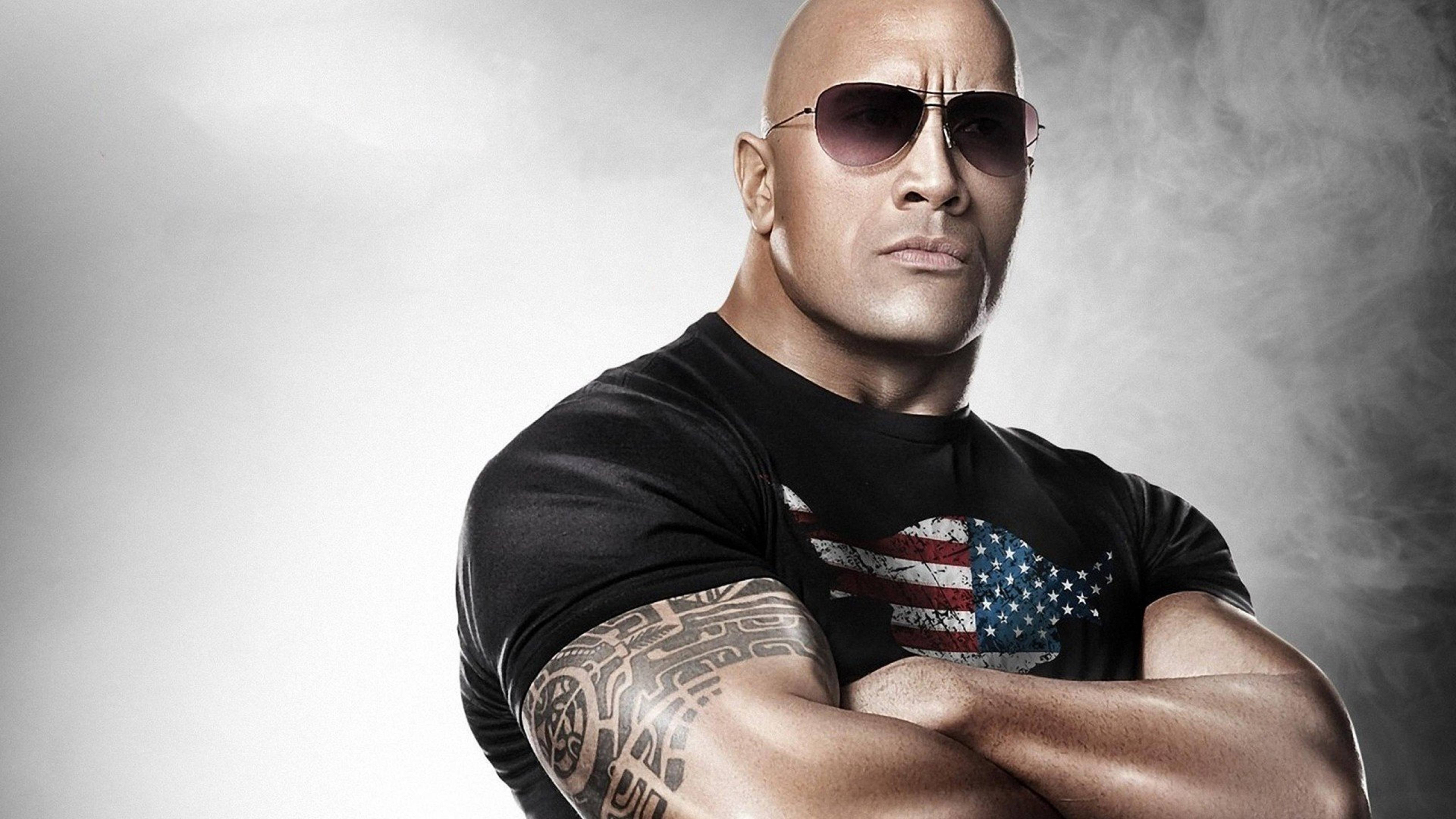 道恩·強森 The Rock 壁紙 — 狄維·莊遜自信地交叉雙臂站立，戴著太陽眼鏡，身穿帶有美國國旗圖案的黑色T恤，背景是煙霧灰色。