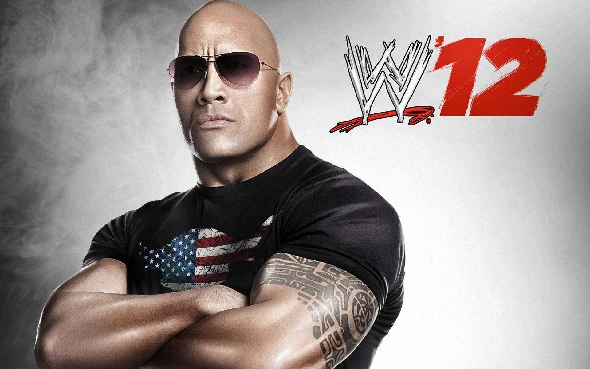 道恩·強森 WWE 12 壁紙 — Dwayne Johnson 戴上太陽眼鏡和美國國旗T恤，雙臂交叉，自信地站在煙霧灰色背景前，帶有WWE 12標誌。