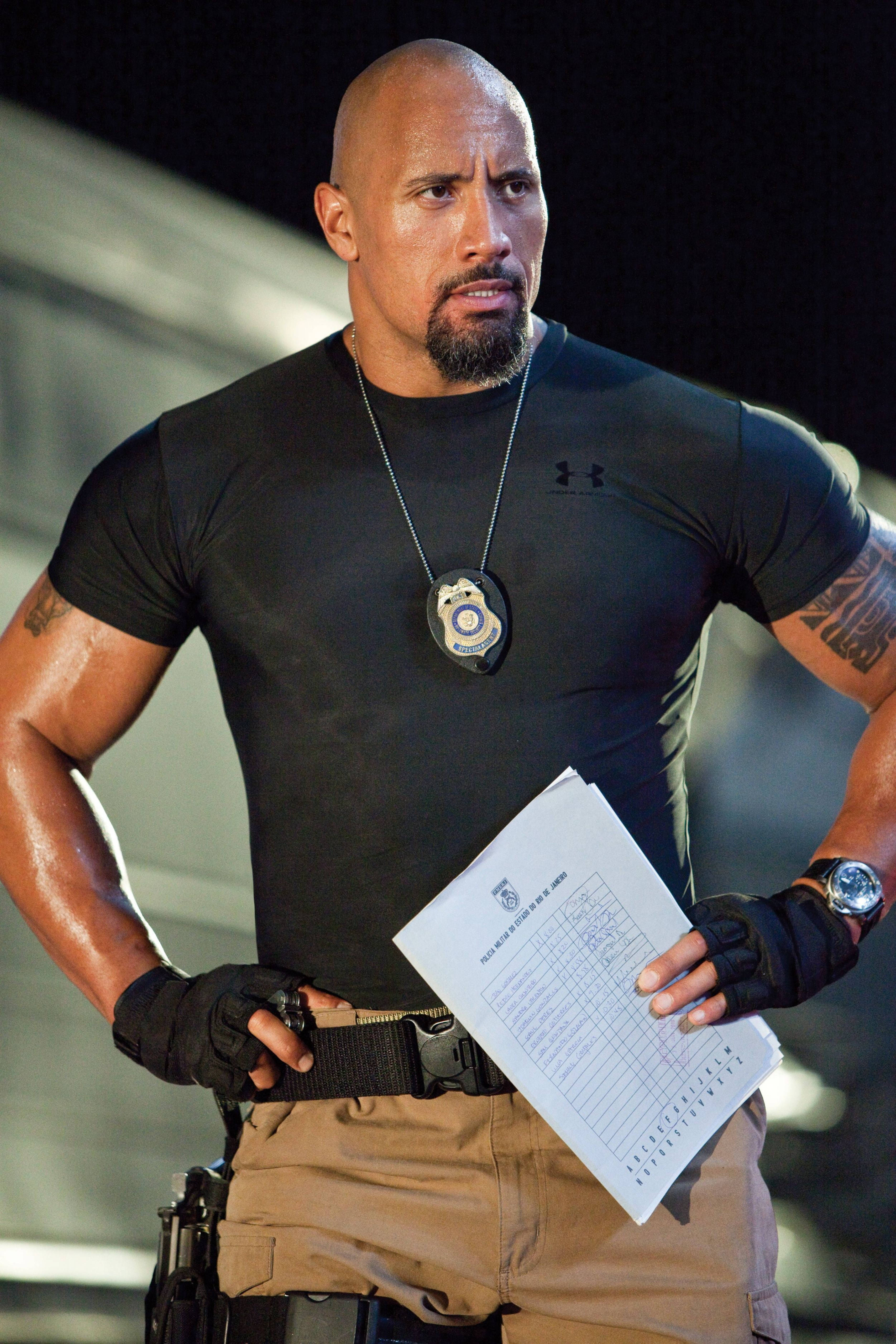 道恩·強森在《玩命關頭5》中 壁紙 — Dwayne Johnson 飾演一名肌肉發達的警察，身穿黑色襯衫，手持報告，表情嚴肅。