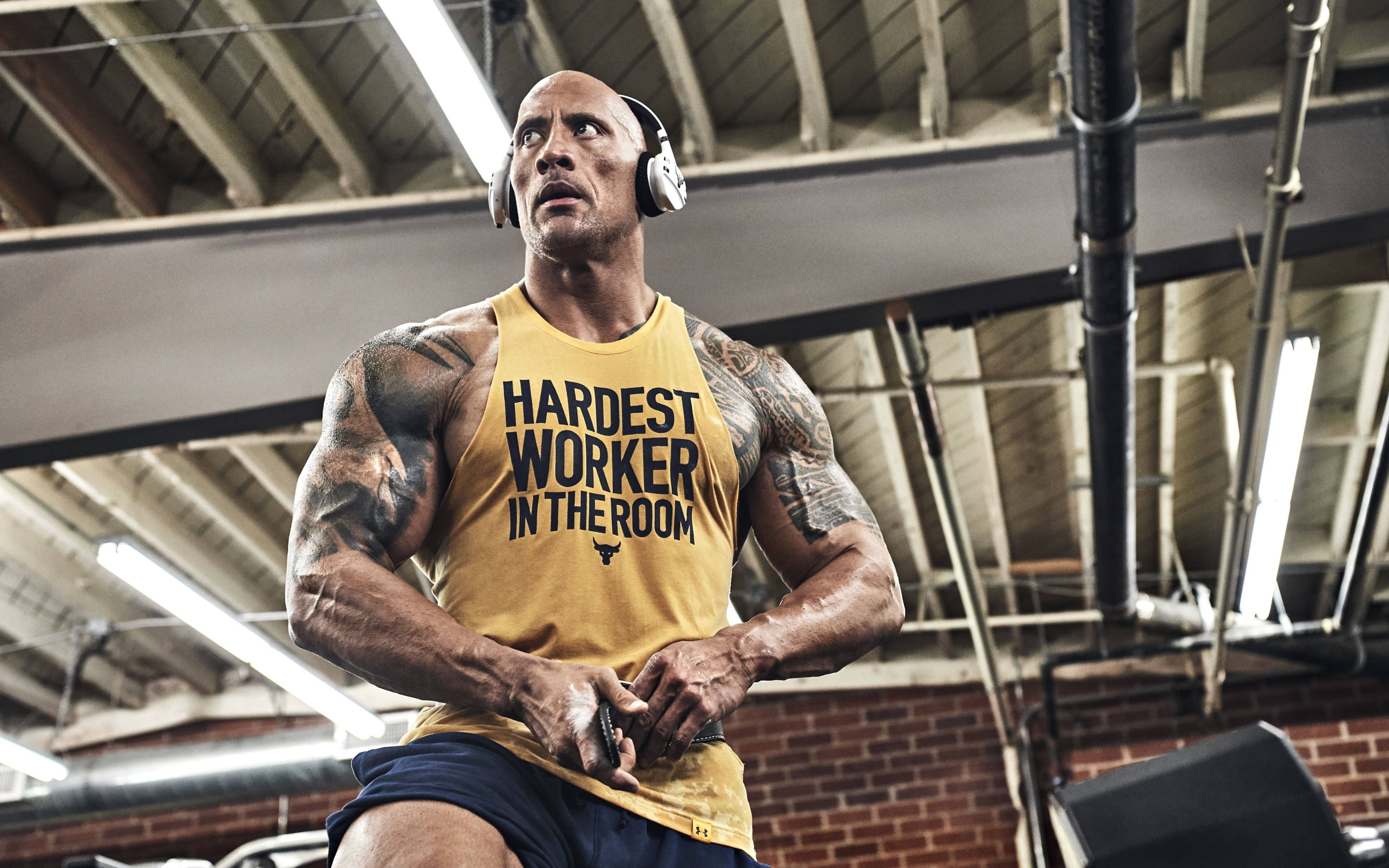道恩·強森宣傳活動 壁紙 — Dwayne Johnson 在健身房戴著耳機，身穿印有 'Hardest Worker in the Room' 字樣的黃色背心。
