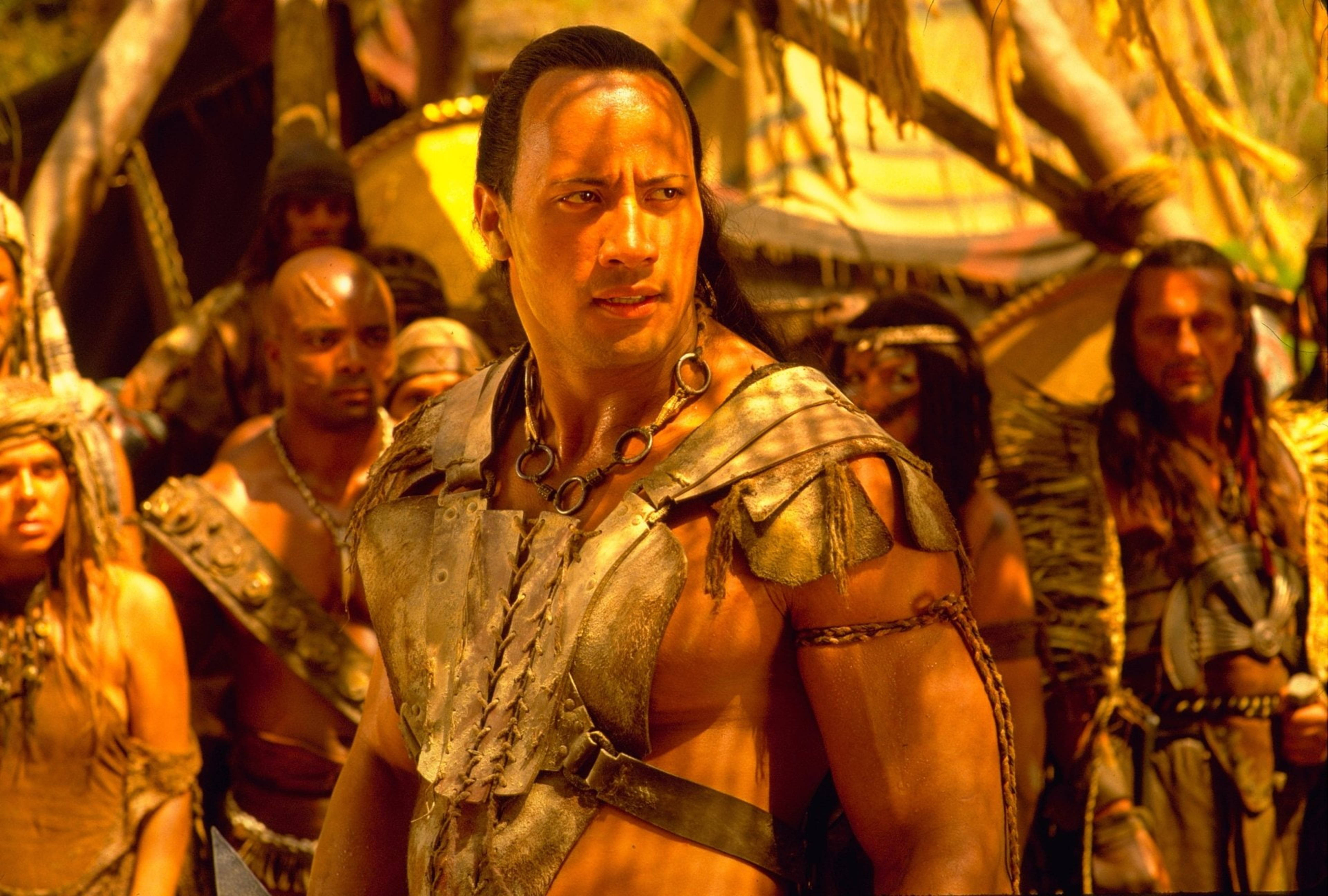 道恩·強森飾演傳奇「蠍子王」 壁紙 — Dwayne Johnson 飾演一位肌肉發達的戰士，身穿部落盔甲，站在畫面中央，周圍是叢林環境中的其他戰士。