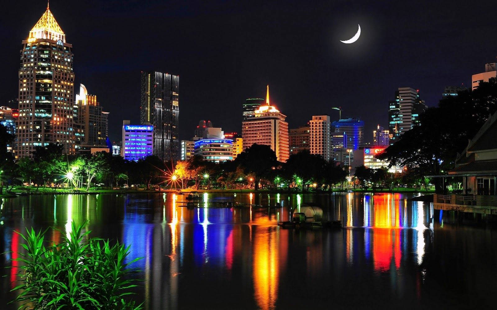 達卡夜景：璀璨建築 壁紙 — 新月下，繁華的城市天際線在平靜的水面上倒映，建築物閃爍著光芒，綠意盎然。